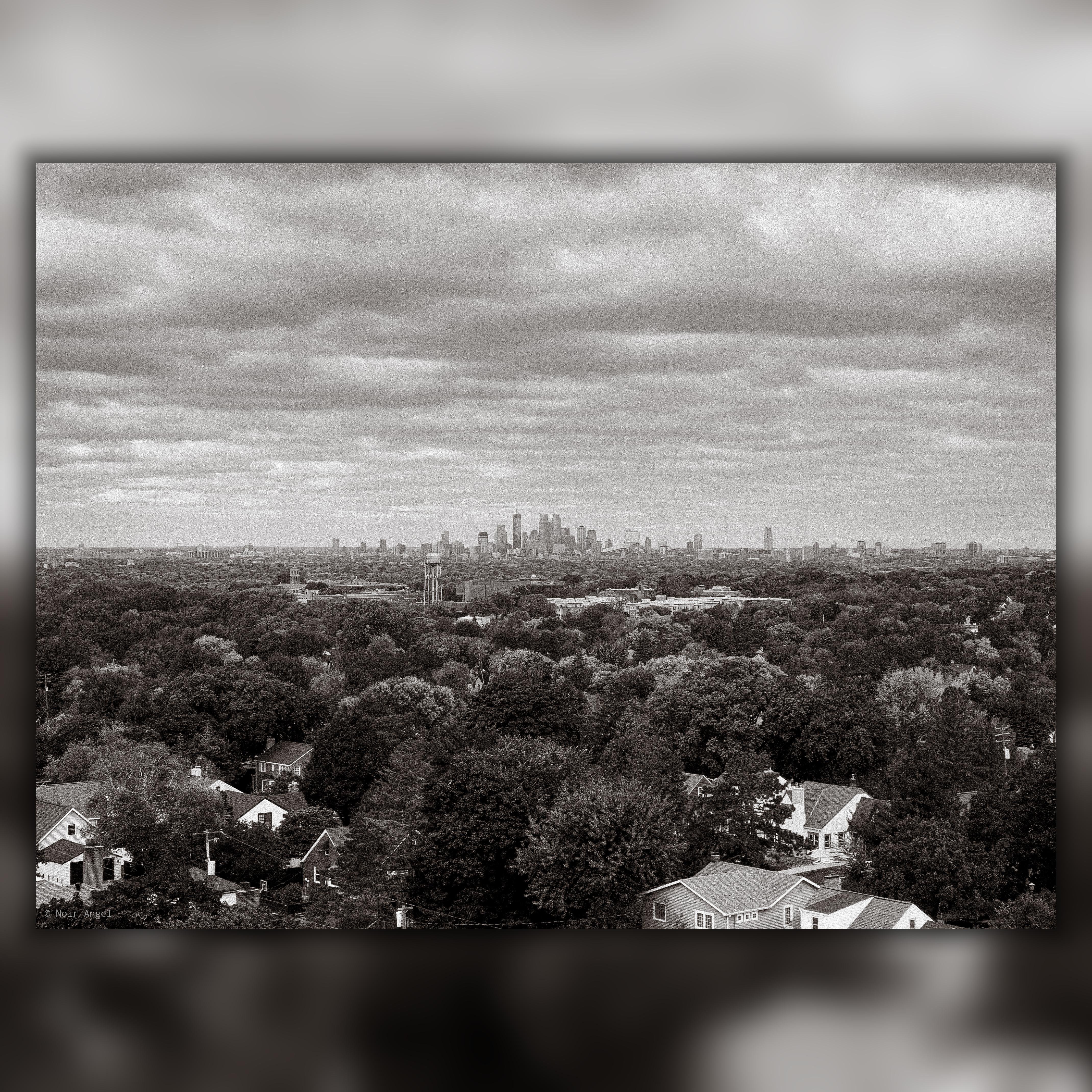 Black and white aerial view of a cityscape with a foreground of densely packed trees and houses. The skyline of a city with tall buildings is visible in the distance under a sky filled with clouds.