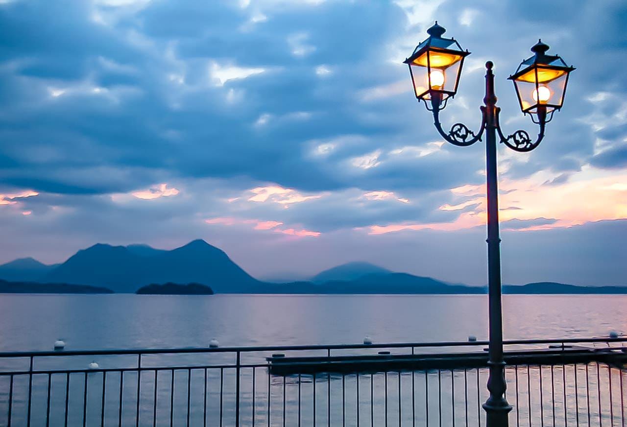 Two-lantern streetlight glowing beside a lakeside railing at dusk, with calm water and silhouetted mountains under a cloudy blue-and-pink sky.
