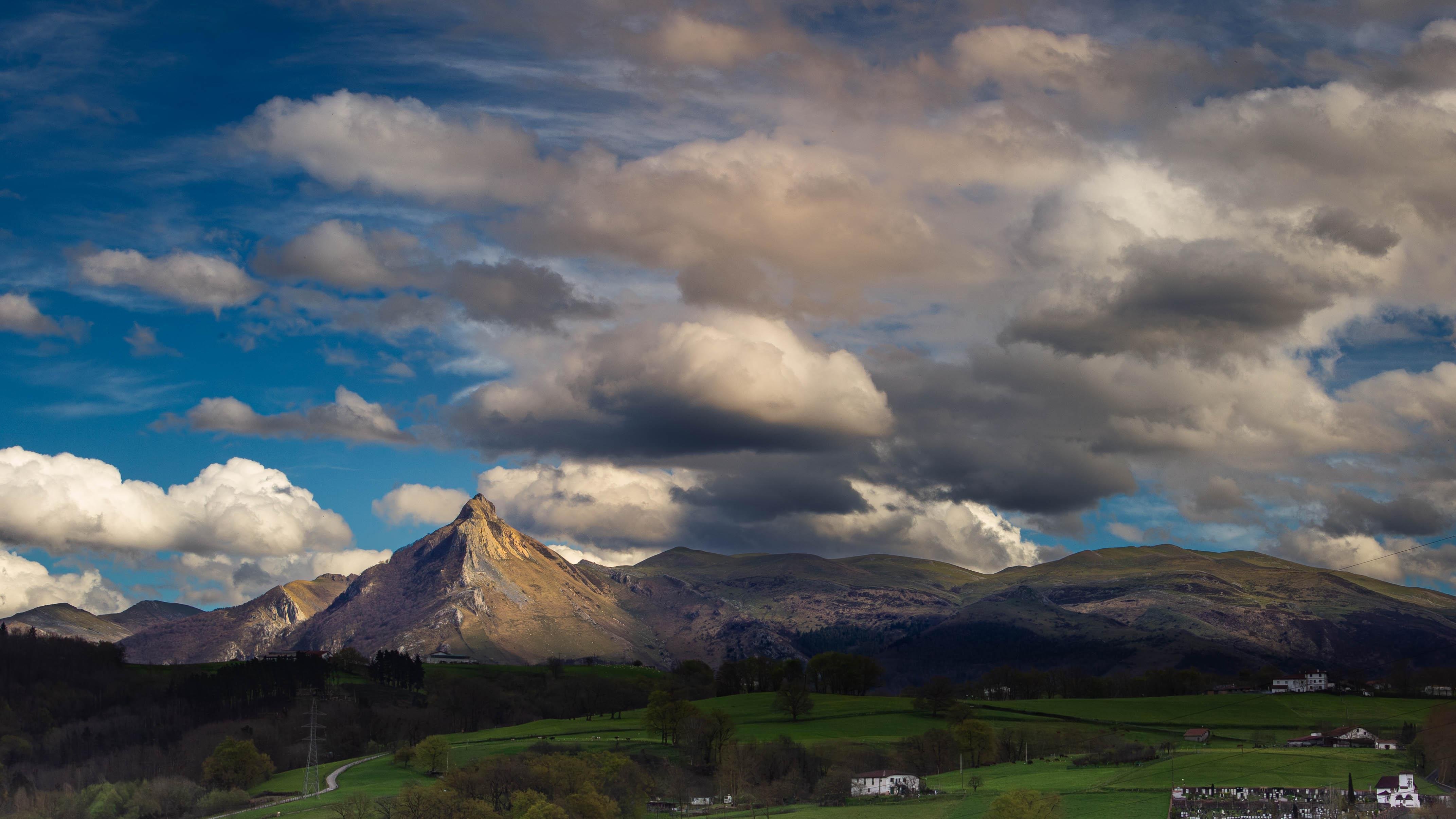 The image features a scenic landscape with a prominent mountain range under a partly cloudy sky. The foreground consists of rolling green hills with some trees, and there are a few buildings visible, likely rural houses or farms. The lighting on the mountains suggests it could be late afternoon, with a mixture of sunlight and shadow adding depth to the scene.