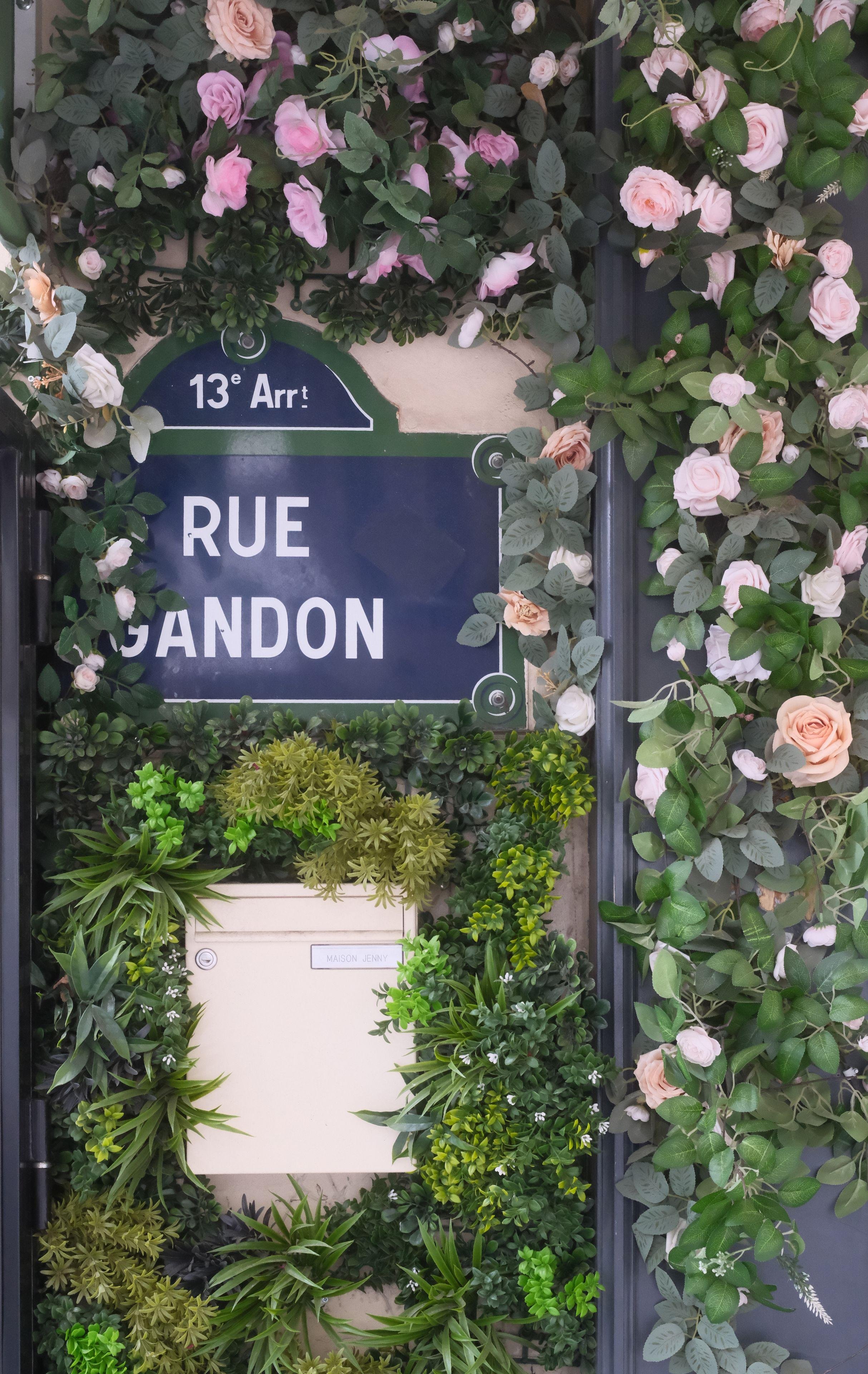 Photo verticale. Une plaque de nom de rue et une boîte aux lettres un peu cachées par des fausses plantes et fleurs accrochés au mur