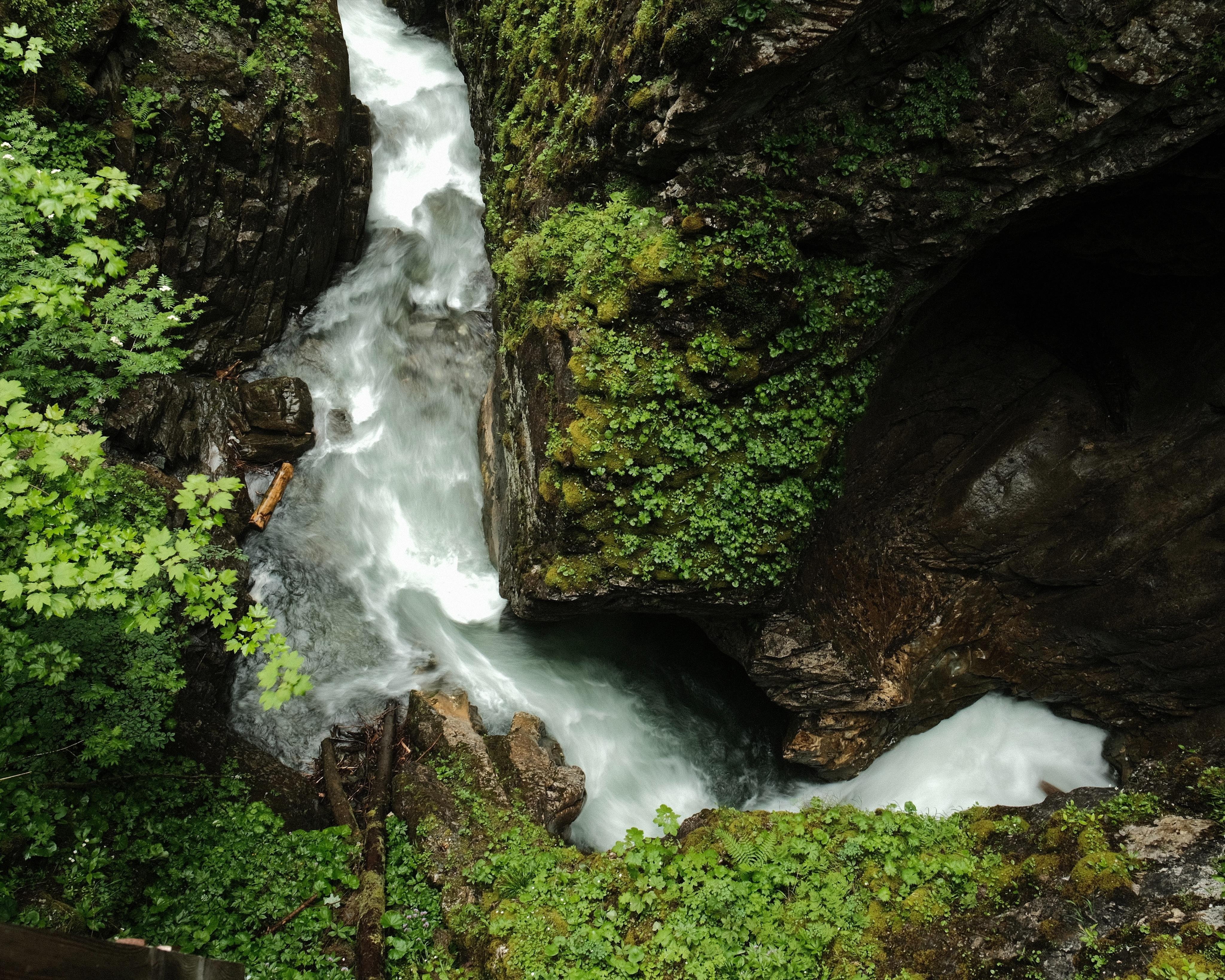 Aerial photo of a narrow gorge with a wild stream flowing between plant-covered rocks. The gorge curves from the right centre to the lower left and then up to the  upper left.
The relatively long exposure generates strong, dynamic lines along the water flow without smoothing the water too much, contrasting the sharp and detailed small leaves and moss on the rocks.