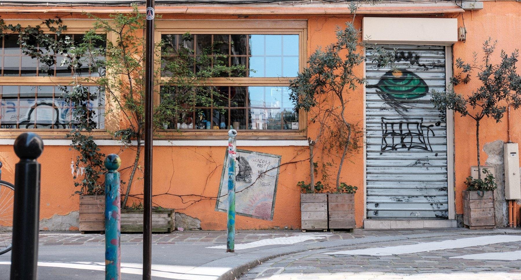 Photo horizontale. Façade du rez de chaussé d’un bâtiment, de couleur orange, des fenêtres dont toutes les demarcations sont aussi oranges. Une porte protégée par un rideau de fer fermé qui est taggé avec un personnage vert et des lettres noires.