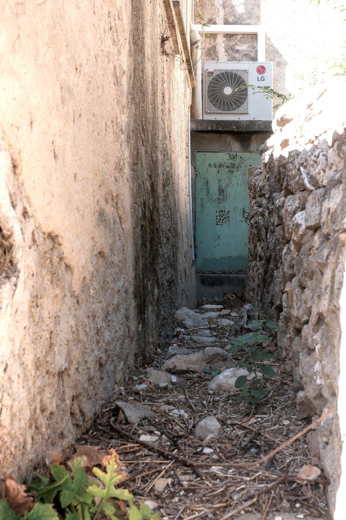Vertical photo. A footpath that looks not quite maintained, with a lot of vegetal debris, at the end of which a metalic green door under an external AC unit. The whole place gives a no man’s land vibe