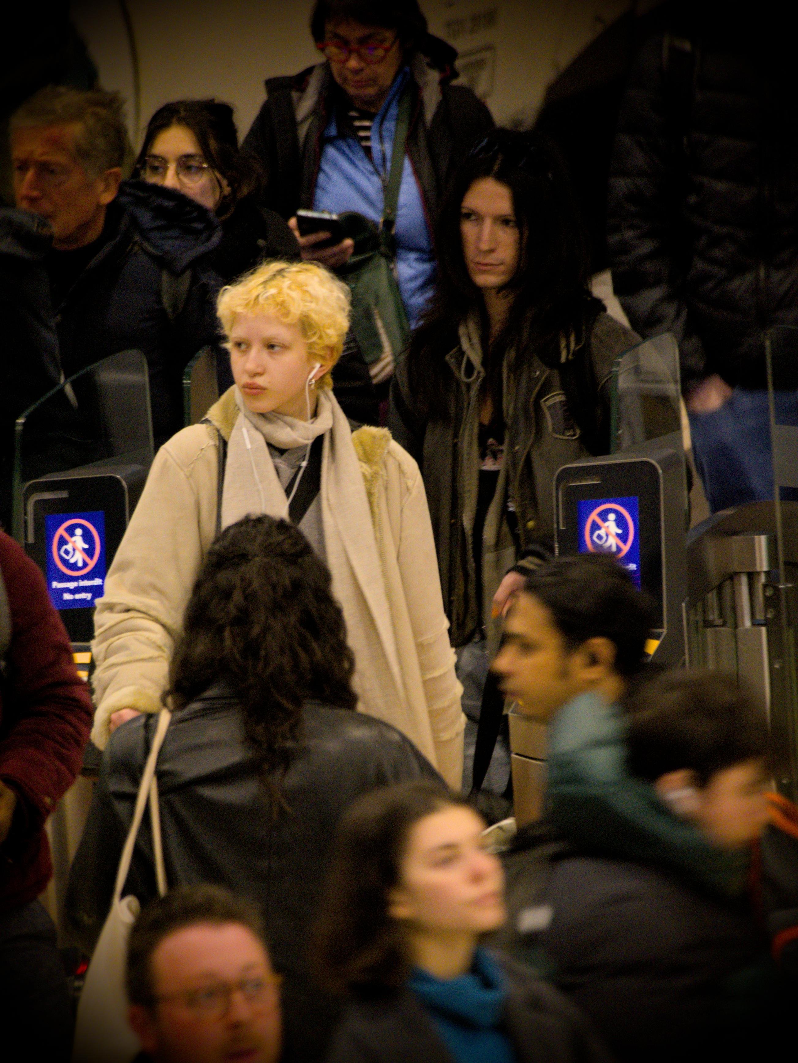La photographie représente une foule animée dans une gare. Les gens se déplacent dans différentes directions, et certains portent des manteaux et des écharpes. Au centre de l'image, alors que tout le monde est habillé de couleurs sombres, une personne ressort en étant de peau très claire, aux cheveux blonds et habillé d'un beige clair.