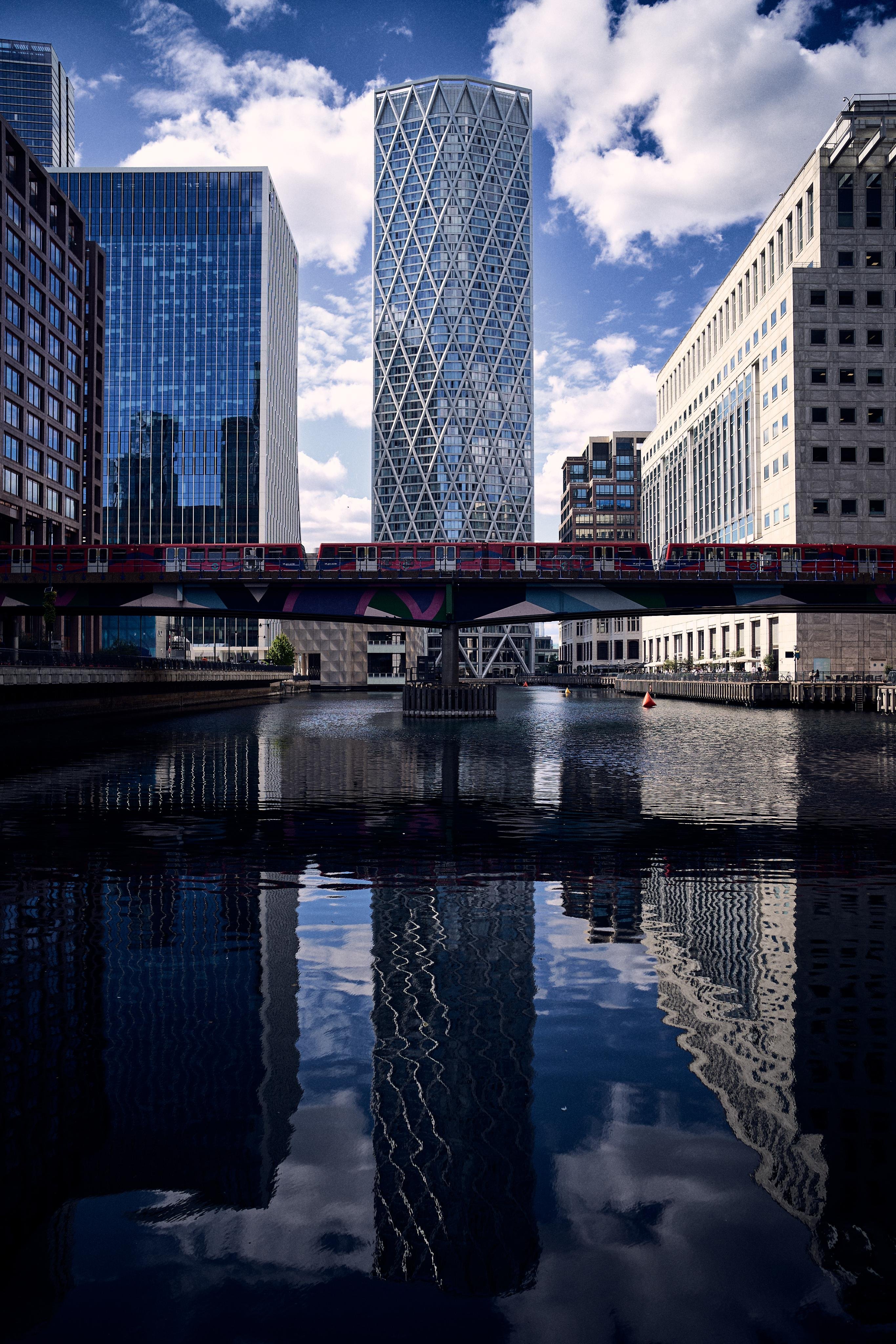 The image depicts a cityscape with modern skyscrapers reflecting in the water below. There's a red train crossing a bridge, and the buildings have a mix of glass and geometric designs. The sky is partly cloudy, and the overall setting looks urban and contemporary.