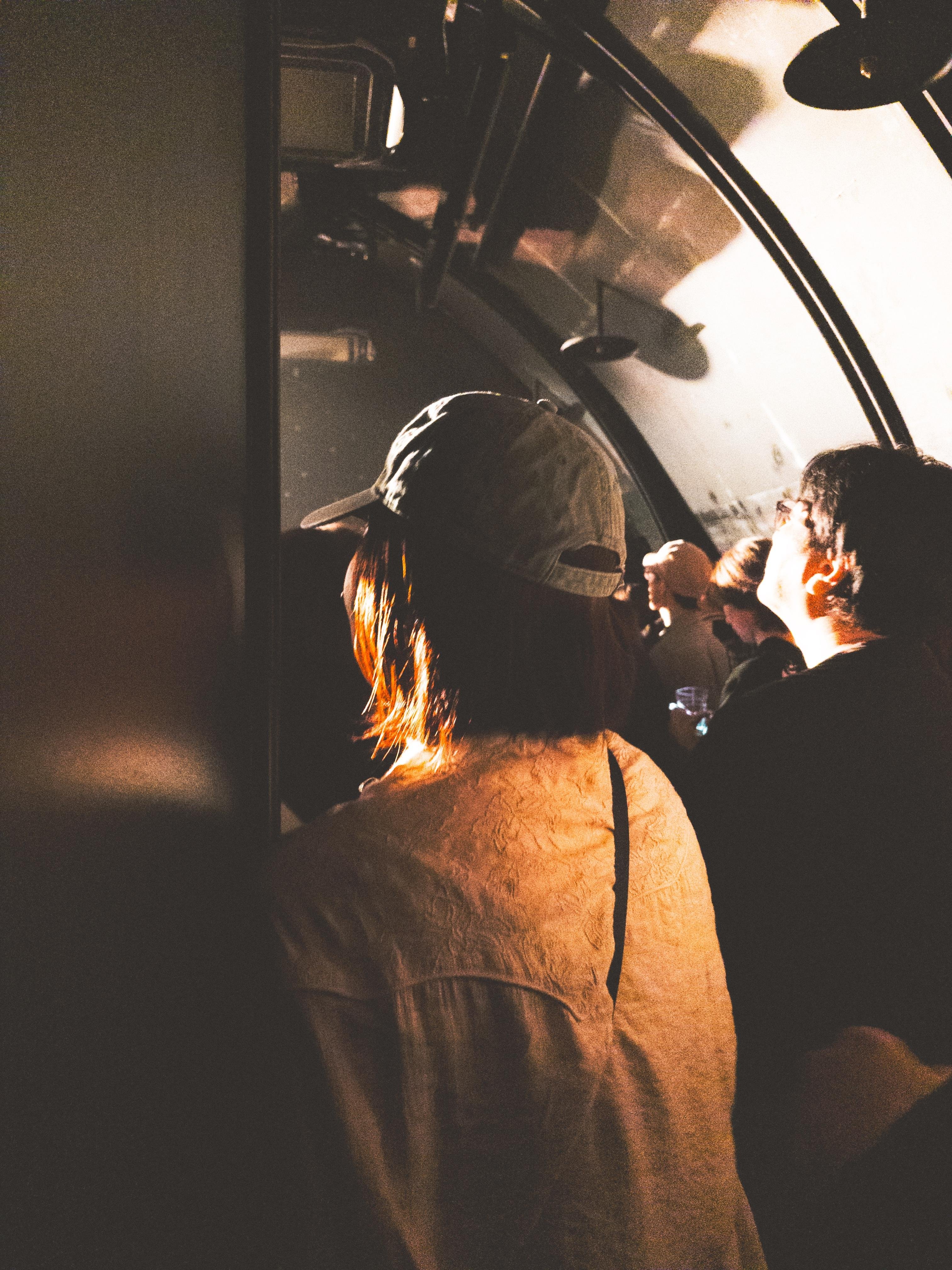 Interior of a music event venue with several people standing closely together, dimly lit by overhead lights. A person in the foreground, wearing a light-colored shirt and a baseball cap, faces forward. The curved metal ceiling and side windows create a tunnel-like atmosphere. Shadows play across the scene, highlighting the passengers' profiles.