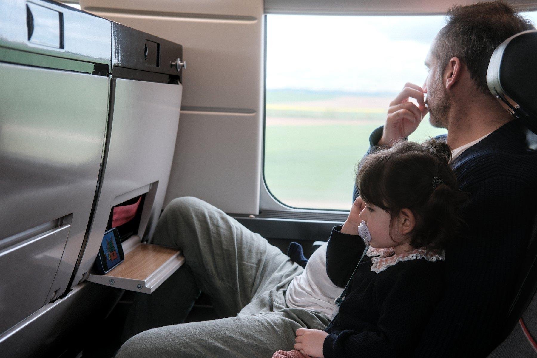 Photo horizontale. Dans un train, une fillette regarde un dessin-animé sur un iPhone en jouant négligemment avec ses cheveux brun de la main droite, aux côtés de son père, qui regarde par la fenêtre