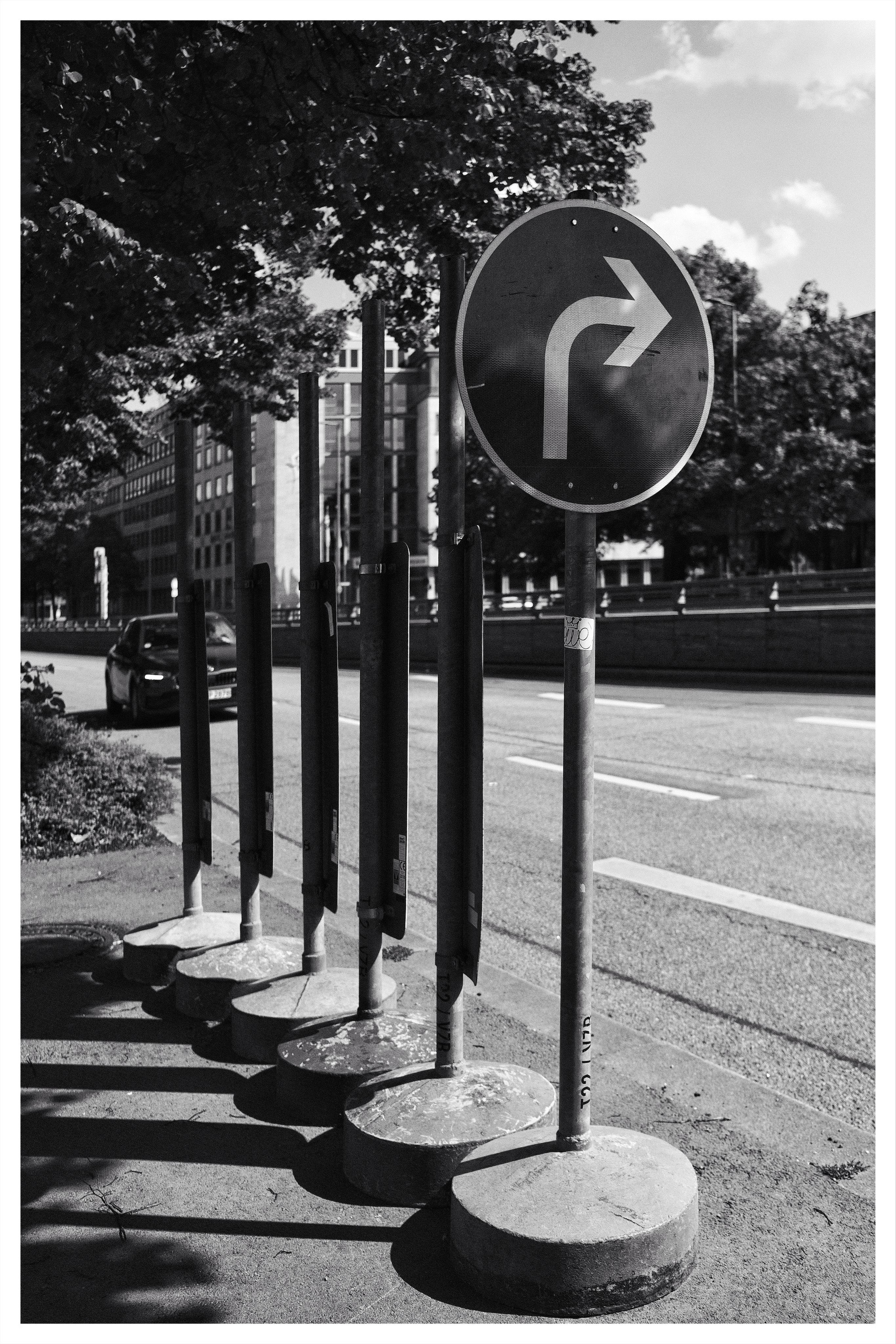Black-and-white street scene with a circular traffic sign showing a right-turn arrow beside a row of temporary metal posts on round concrete bases along the sidewalk; a car and multi-story buildings in the background under trees.