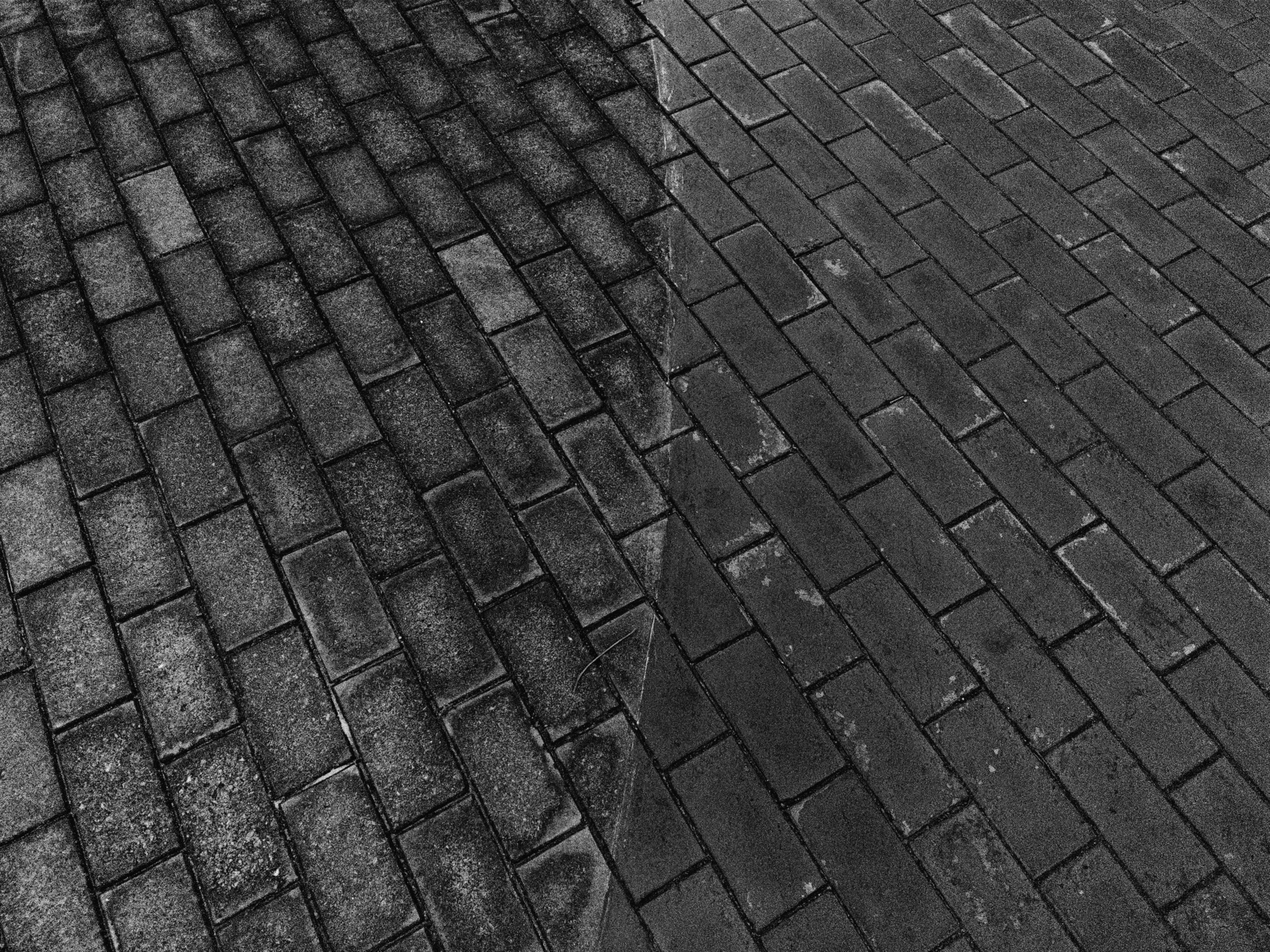 Brick pavement split into two sections, left side showing older, weathered bricks with visible stains and discoloration, and right side featuring newer, cleaner bricks. The two sections meet with a straight dividing line down the center.