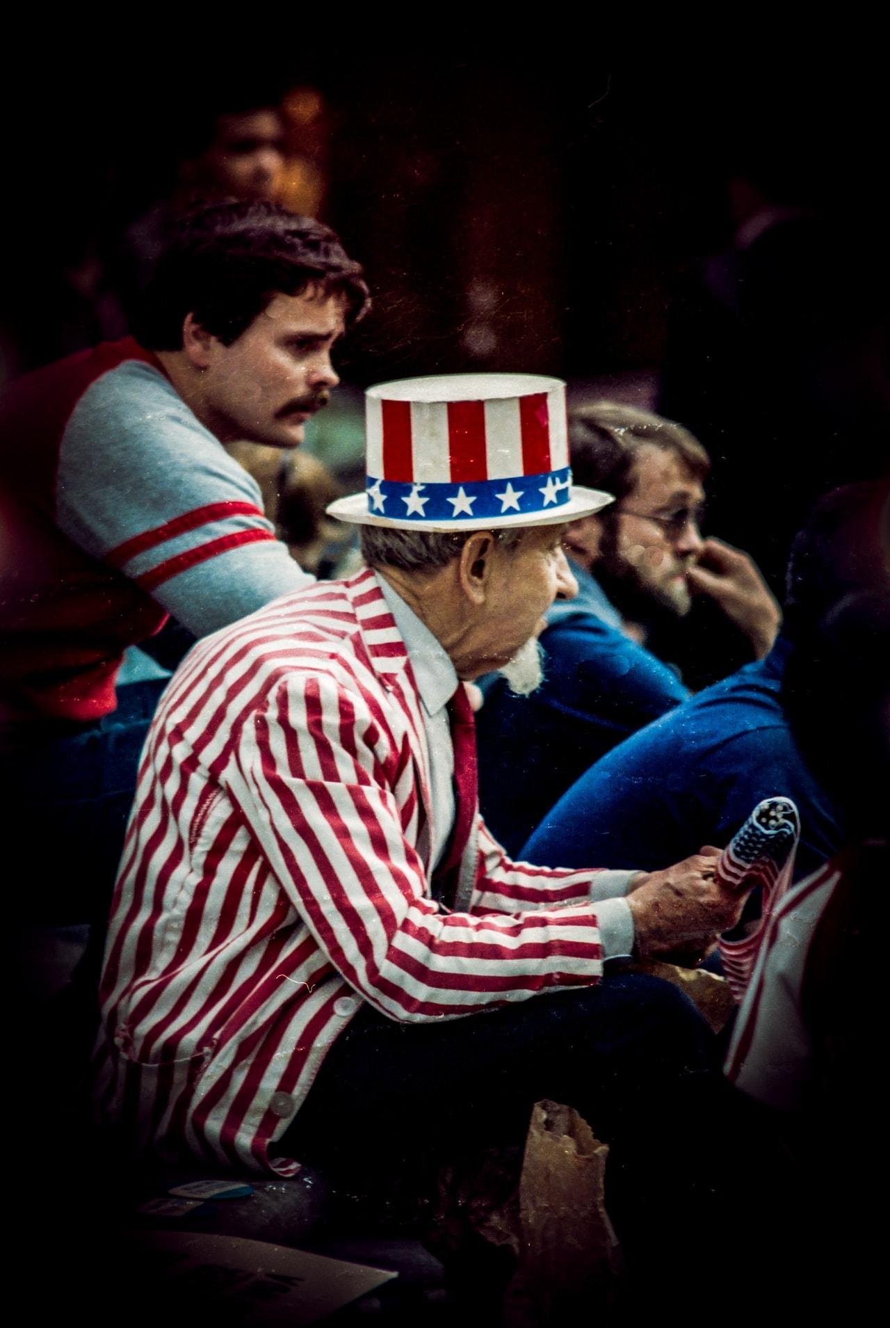 Elderly man wearing a red and white striped jacket and a top hat with an American flag design. He holds a small American flag. Surrounding him are several people seated, some with facial hair, in casual clothing. The background is dimly lit, with the focus primarily on the elderly man.