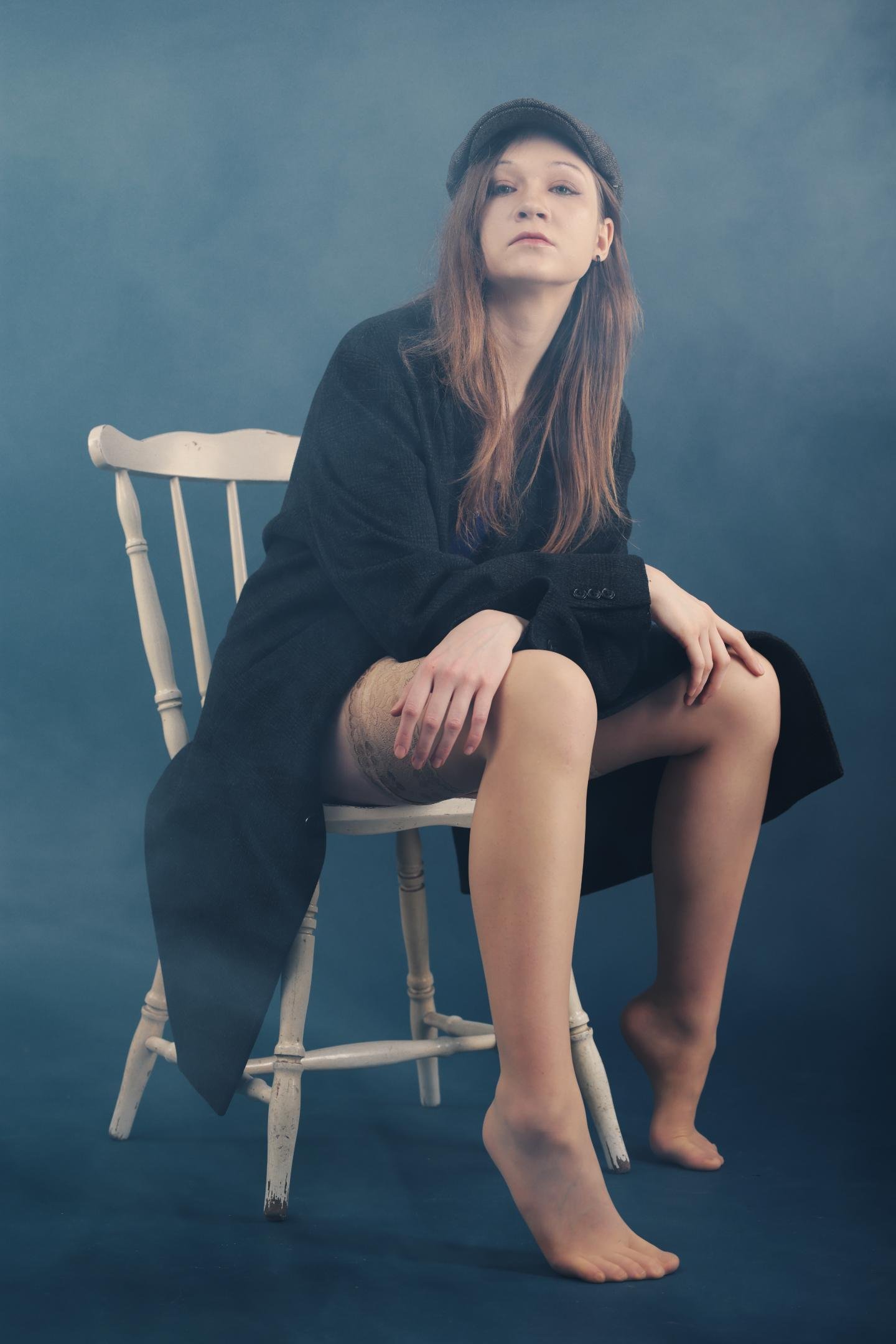 The image shows a person sitting on a wooden chair against a smoky blue background. They are wearing a black coat and a cap, and they are barefoot. The person's expression is neutral or contemplative.
