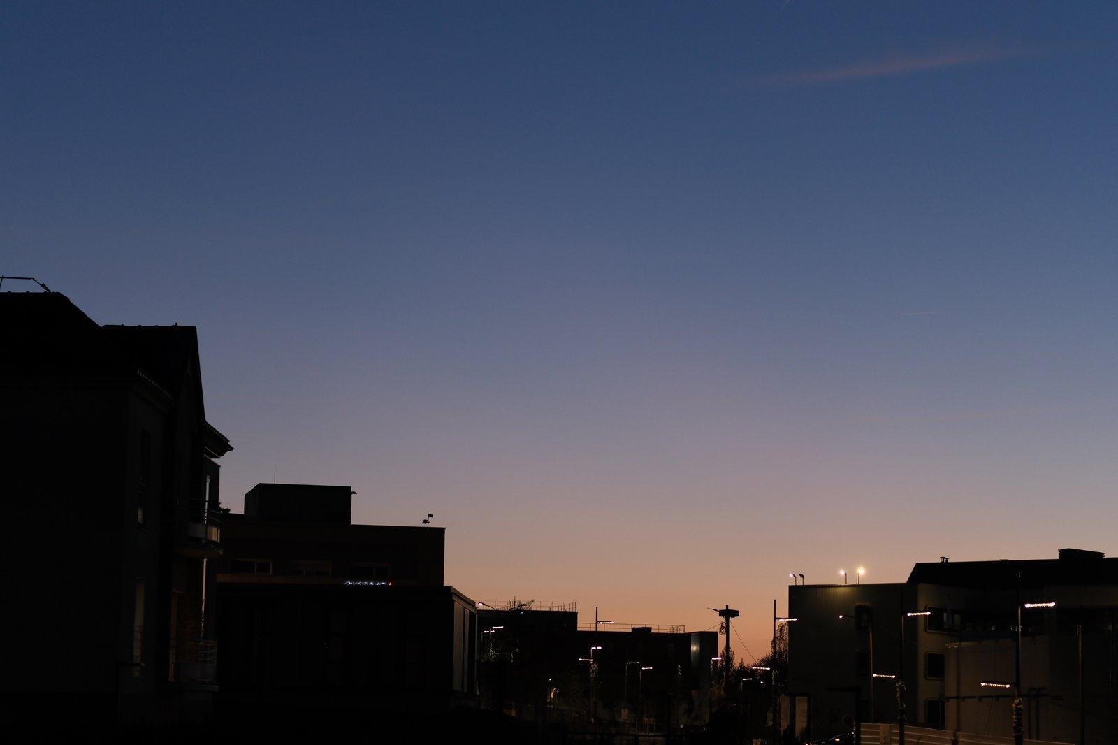Photo format paysage d’un ciel de soleil couchant allant du bleu nuit pastel vers l’orange pastel, au dessus de la silhouette d’immeubles en arc de cercle dans l’image
