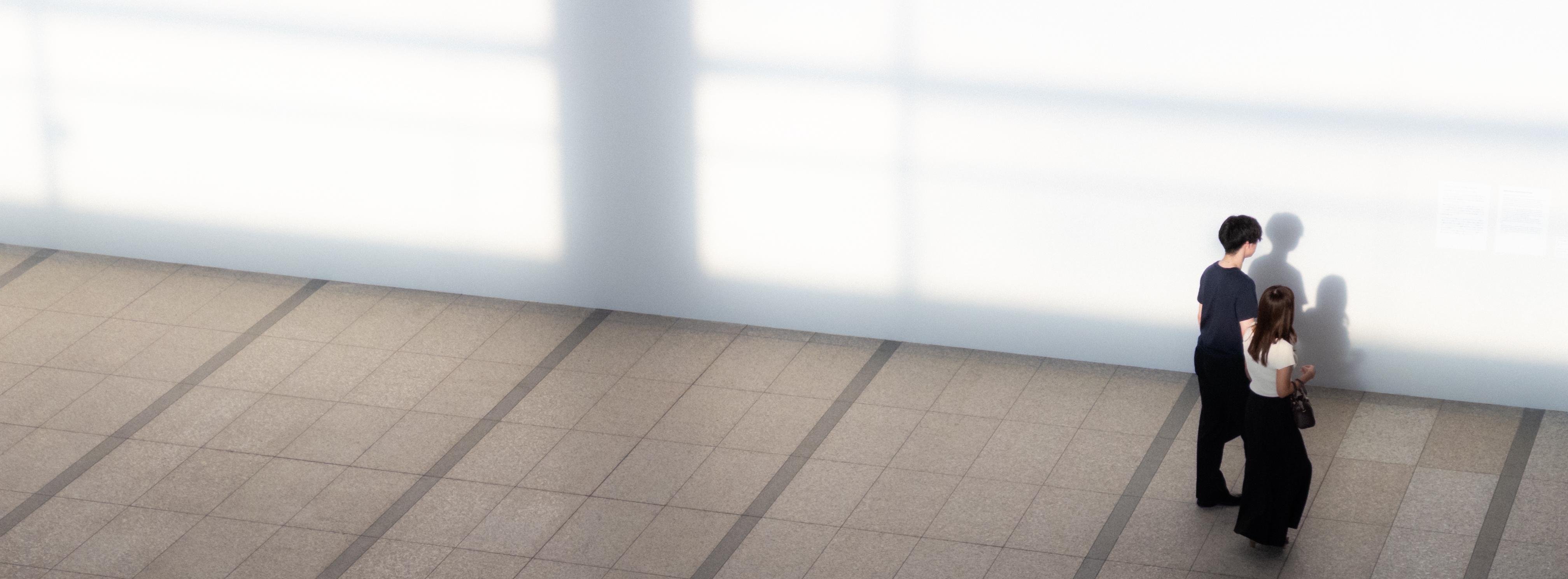 Two people walking side by side along a spacious, tiled floor with a large, white wall on the right casting soft shadows; the ground displays a grid pattern of gray tiles with darker lines.