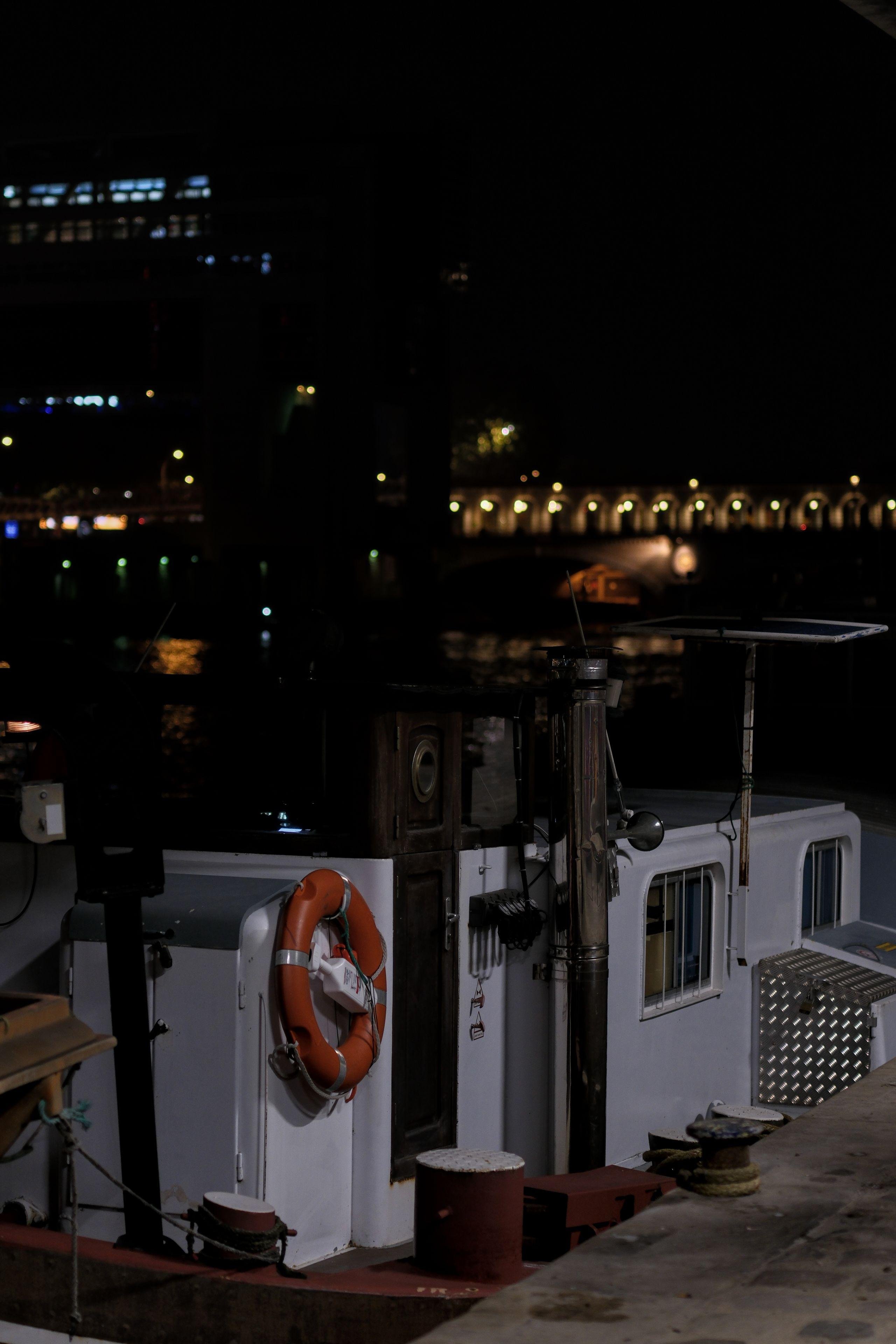 Photo verticale de nuit. Un petit bateau amarré à un quai. Il est blanc avec une bouée de sauvetage rouge accroché au mur de la cabine. Au fond, le noir de la nuit avec une rangée de lampadaires délimitant un pont