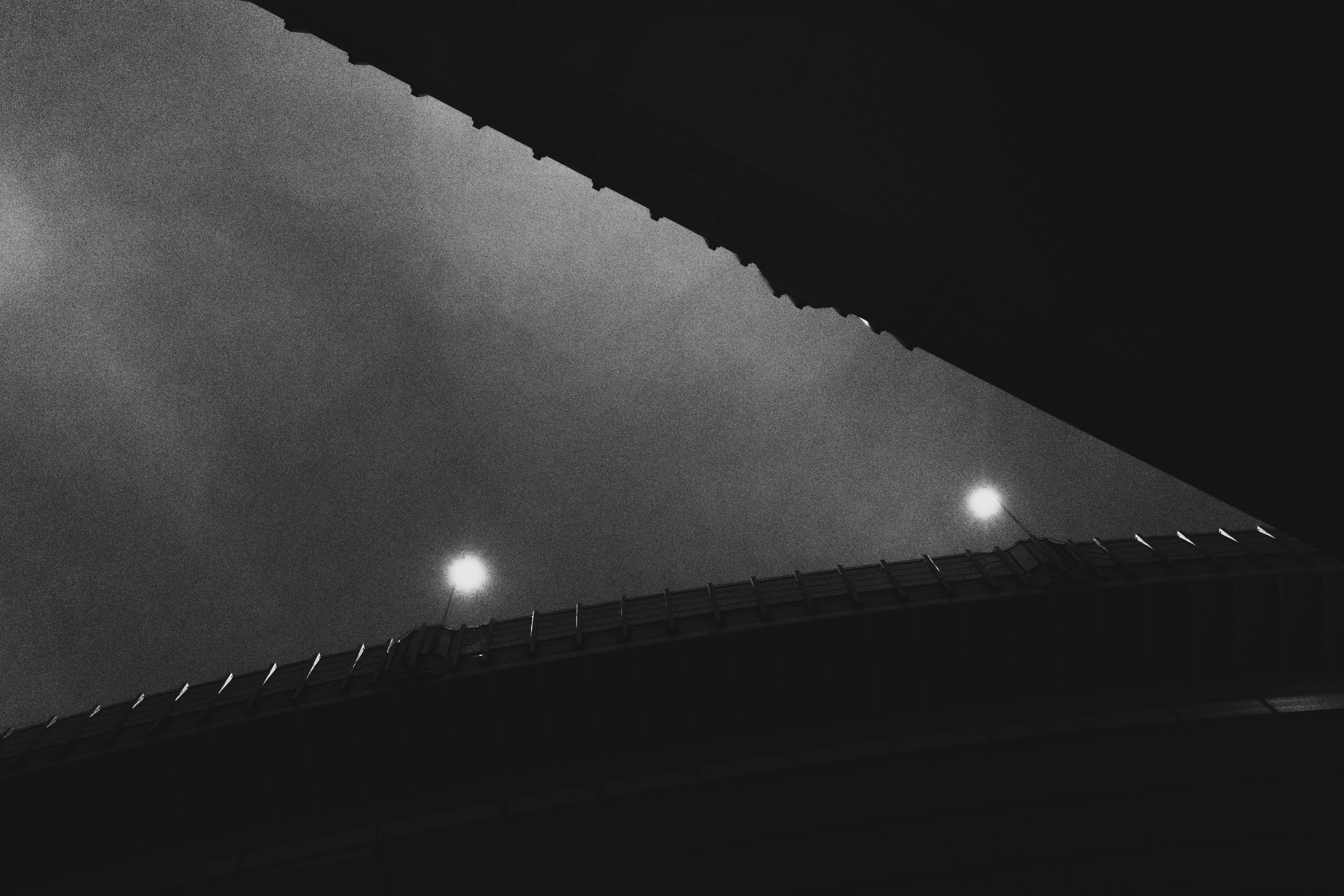 A dark, high-contrast image with a bridge captured from below. The bridge spans diagonally across the middle of the photograph, silhouetted against a cloudy night sky. Two streetlights on the bridge emit soft white light, creating small bright spots in the otherwise dim scene. Edges of the bridge feature a line of repeating geometric patterns.
