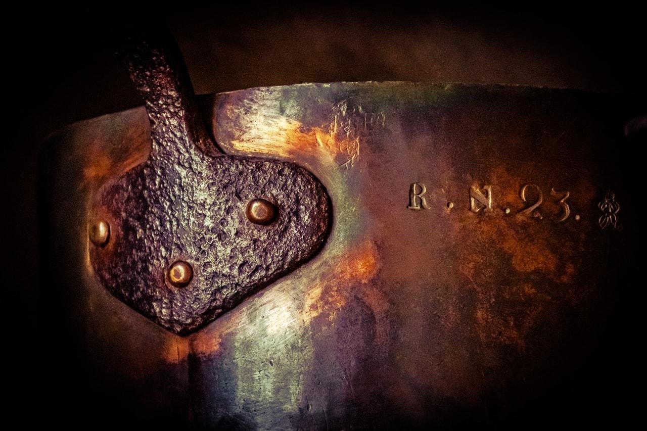Close-up of a worn metal surface with a dark, textured riveted plate on the left and stamped characters “R.N. 23.” on the right, lit with warm highlights against a dark background.