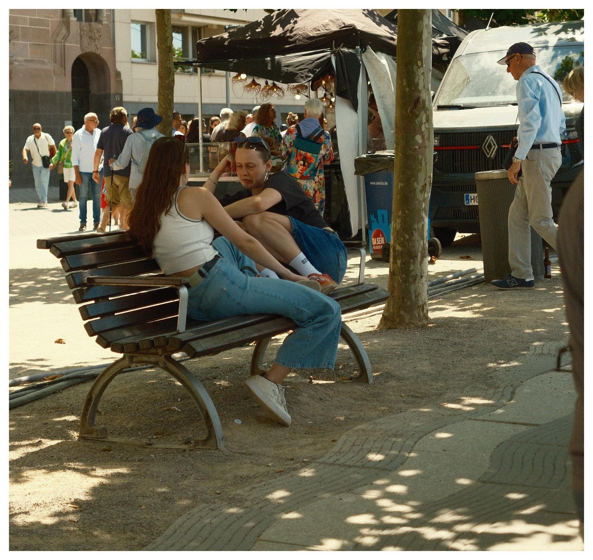 Two people lounging on a wooden park bench under trees, with a busy street market behind them featuring a black canopy tent, a parked van, and pedestrians walking past on a sunlit sidewalk.