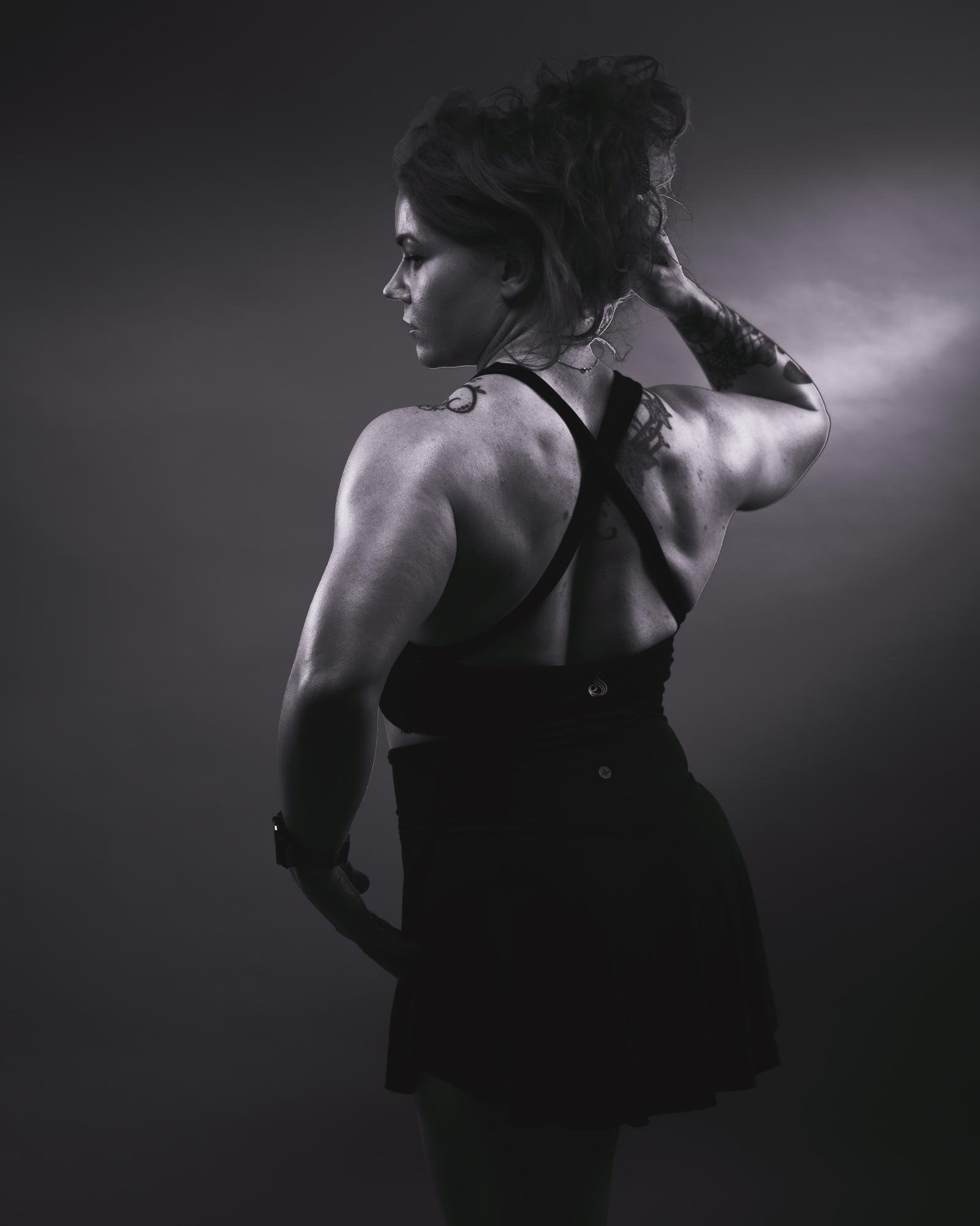 The image is a black-and-white photograph of a person with their back turned, wearing a sleeveless top and skirt. They have a tattoo on one arm and are holding their hair up. The lighting creates a dramatic effect, highlighting the person's muscular build and the texture of their skin.