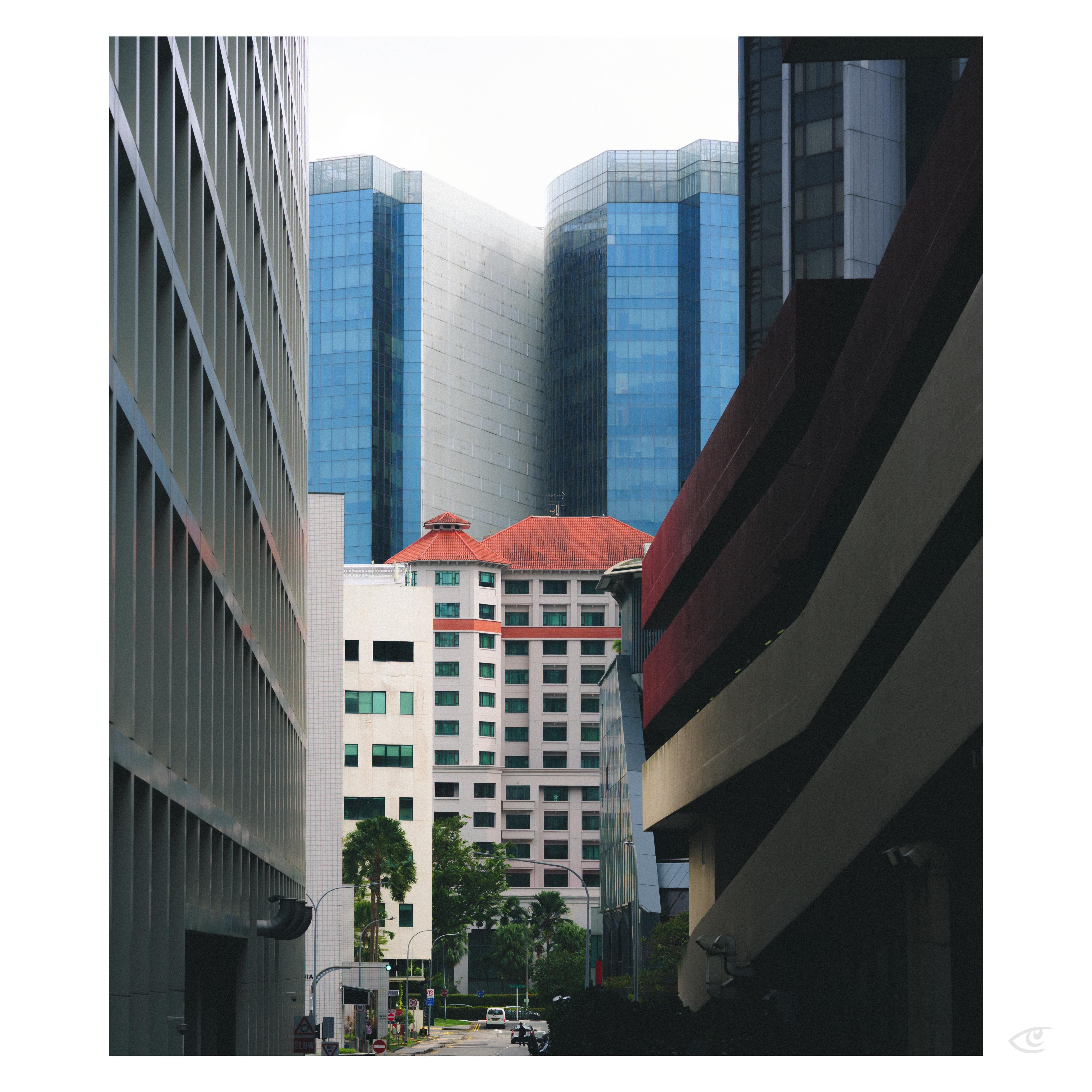 Narrow city street framed by tall buildings on both sides, looking toward a white mid-rise with a red roof and large blue-glass towers rising behind it; small trees, streetlights, and a few vehicles visible at street level.