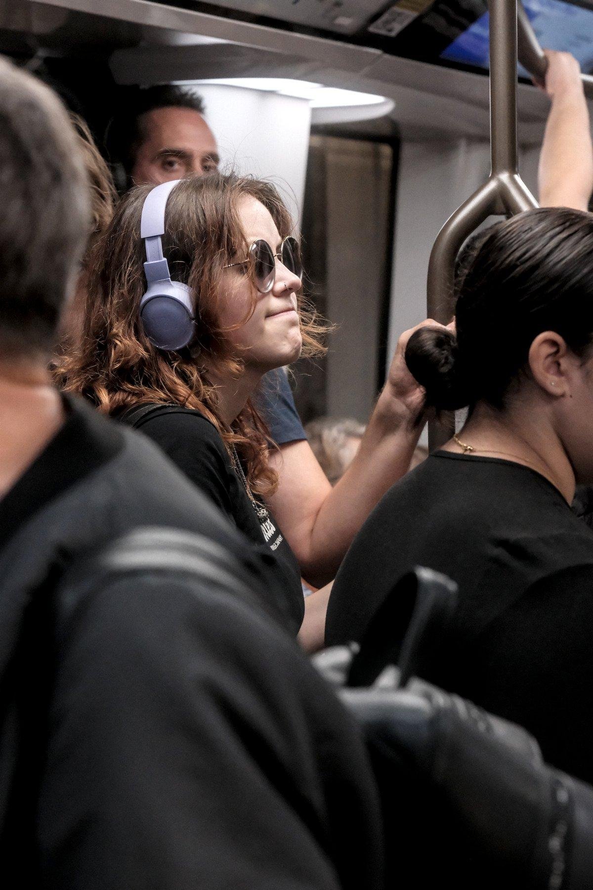 Vertical photo. In a subway, All the clothes are black but the purple headphone of a young woman in the middle of the frame. She’s also wearing sunglasses and is pinching her lips