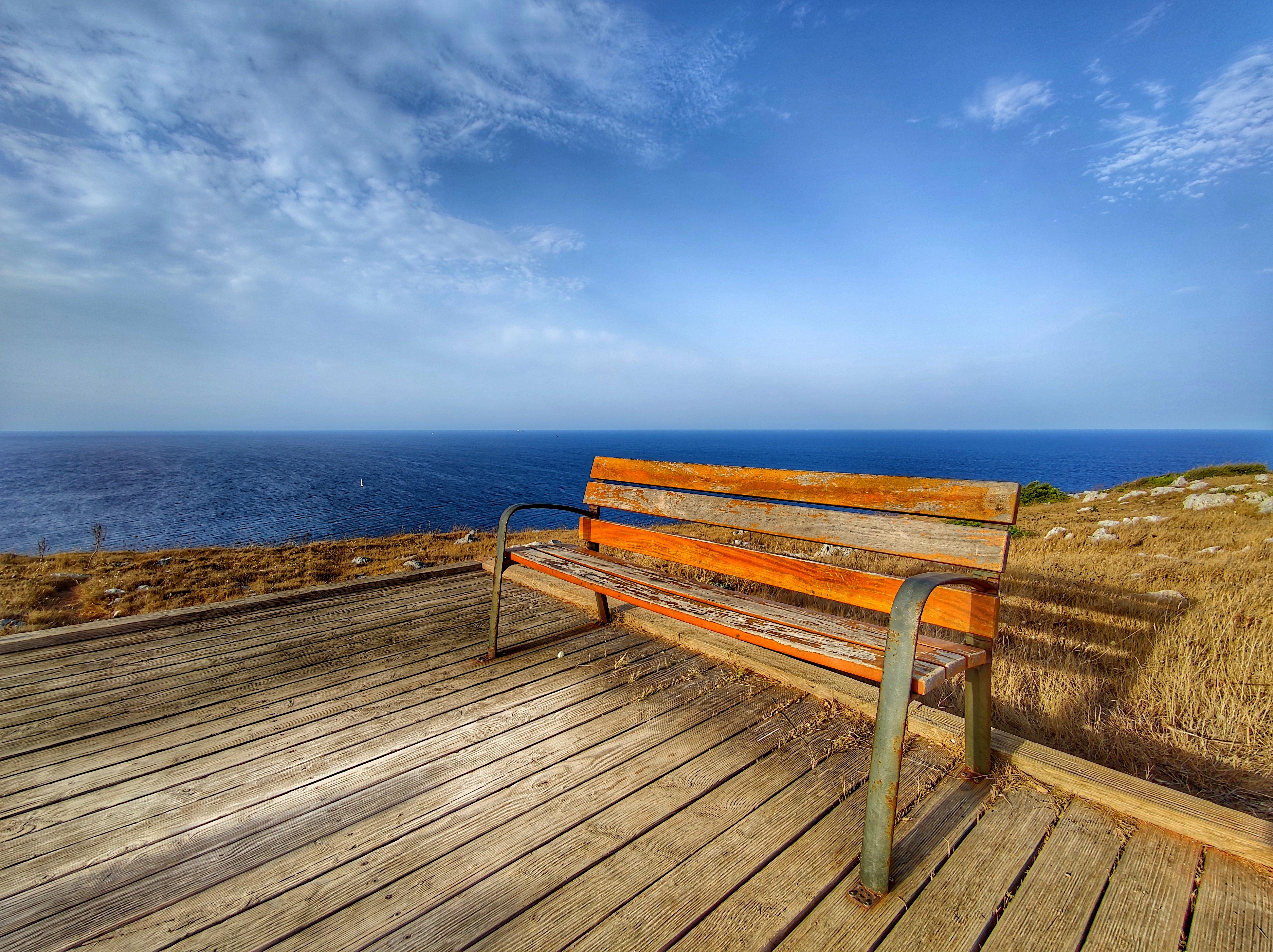 Wooden bench with metal armrests on a wooden deck overlooking the sea. The horizon meets a blue sky scattered with clouds. Dry grass surrounds the deck, which is situated on a coastal landscape.