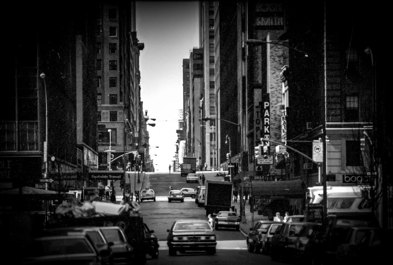 Black and white image of a city street, lined with tall buildings on both sides. Several cars and trucks are visible on the road, heading toward the horizon. Signs and traffic lights are visible, and pedestrians are scattered along the sidewalks. The scene captures a sense of urban density and activity.