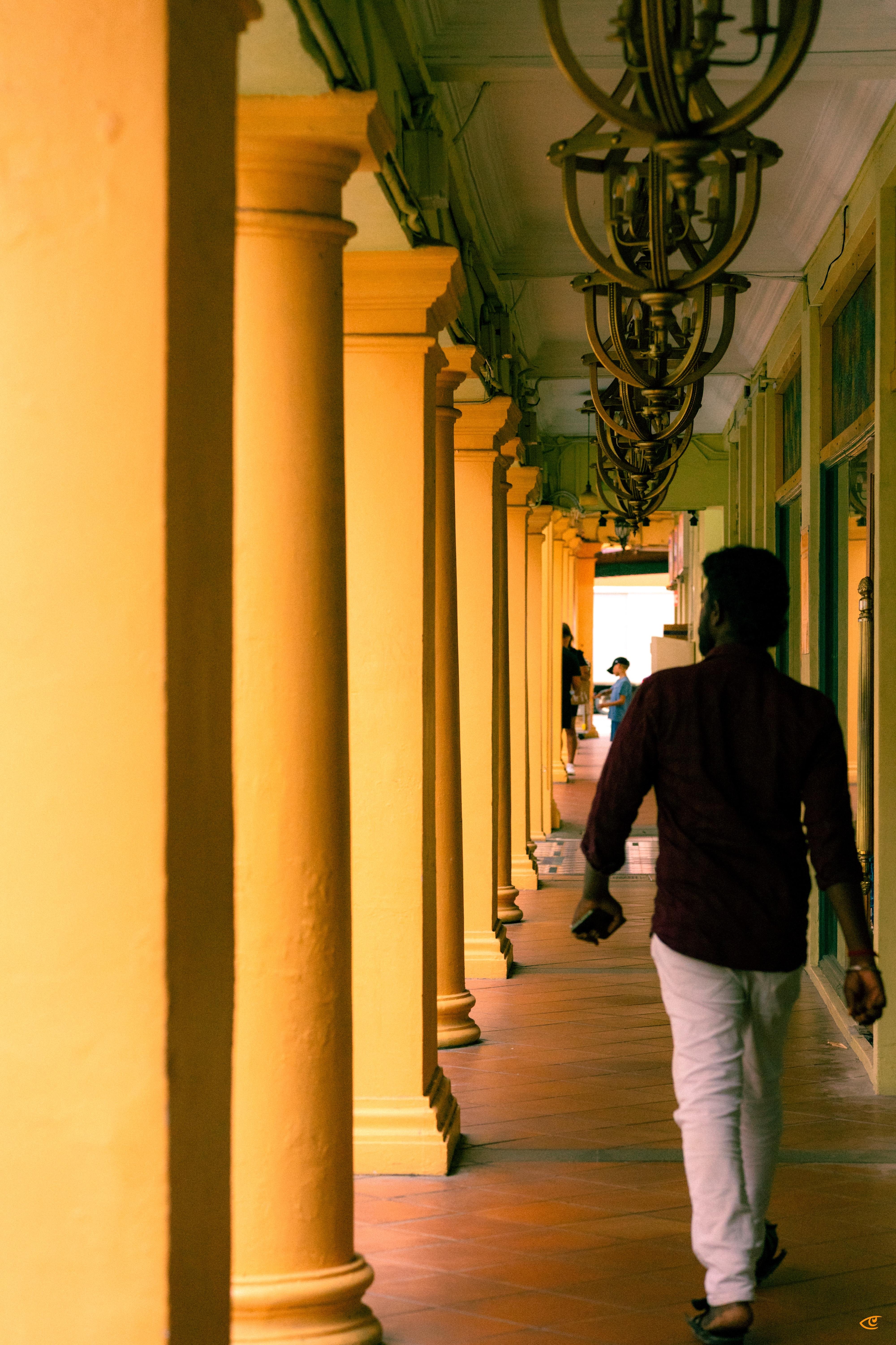 Man walking away down a covered corridor lined with yellow columns, hanging metal lanterns overhead, and storefront windows on the right, with a few people visible in the distance near the bright opening.