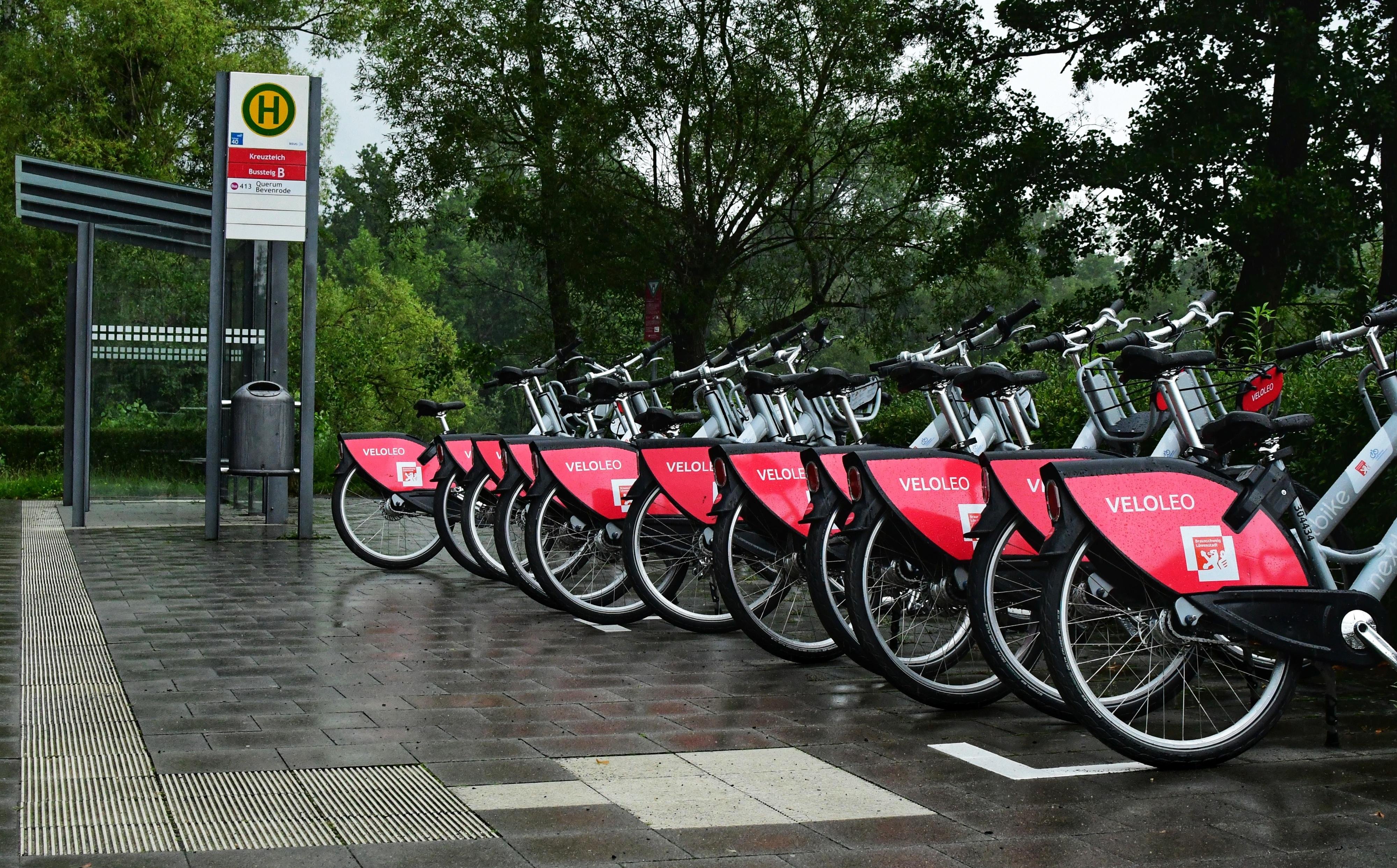 Row of parked rental bicycles with red covers displaying the logo "VELOLEO" on their side stands on a wet paved surface. A nearby bus stop sign indicates public transportation access. Lush green trees form the background under an overcast sky.
###
Eine Reihe geparkter Leihfahrräder mit roten Hüllen und dem Logo „VELOLEO“ steht auf nassem Asphalt. Ein nahegelegenes Bushaltestellenschild weist auf die Anbindung an öffentliche Verkehrsmittel hin. Üppige grüne Bäume bilden den Hintergrund unter einem bewölkten Himmel.