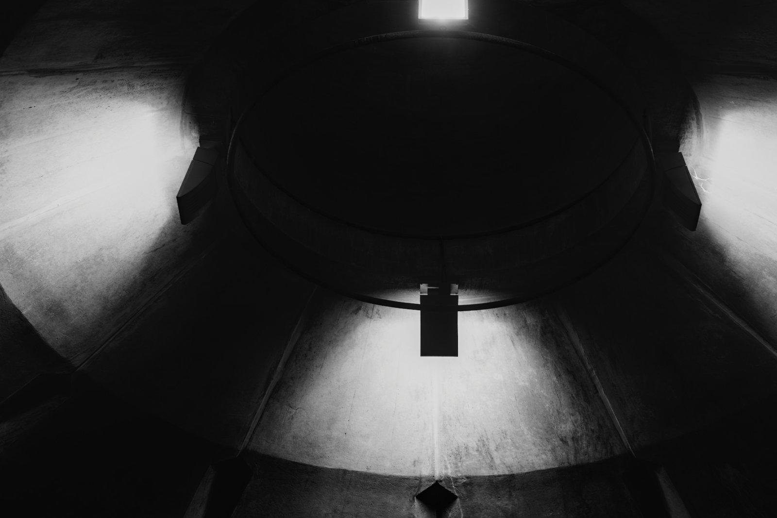 Photo format paysage en noir et blanc, montrant un plafond circulaire avec 4 lampes, dont une hors cadre. Le fort contraste donne une impression d’être dans un vaisseau extra-terrestre