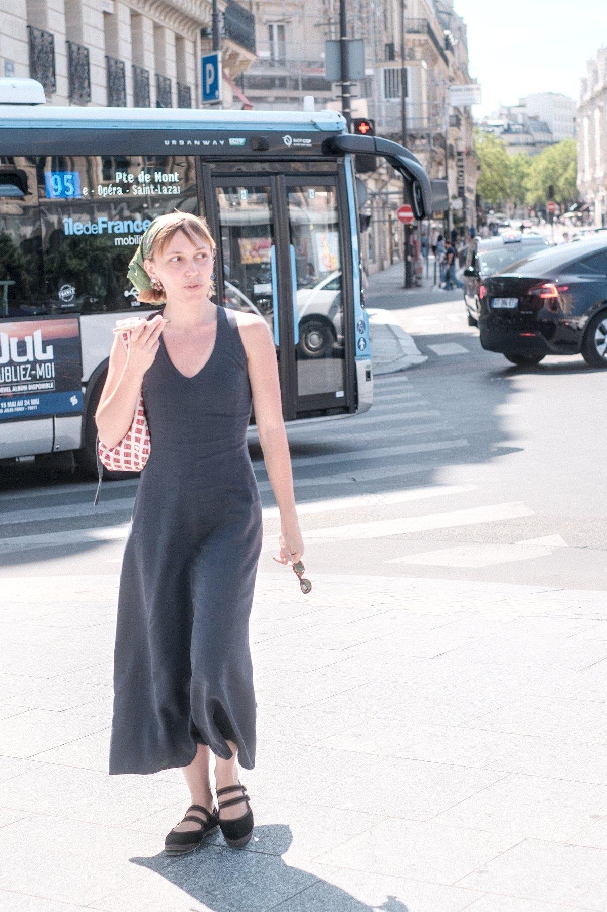 Photo verticale. Une jeune femme en longe robe grise ardoise, avec un bandana vert lui tenant les cheveux roux, entrain de marcher sur un passage piéton en parlant dans son téléphone, en mode haut parleur vu comment elle le tiens au niveau de sa bouche