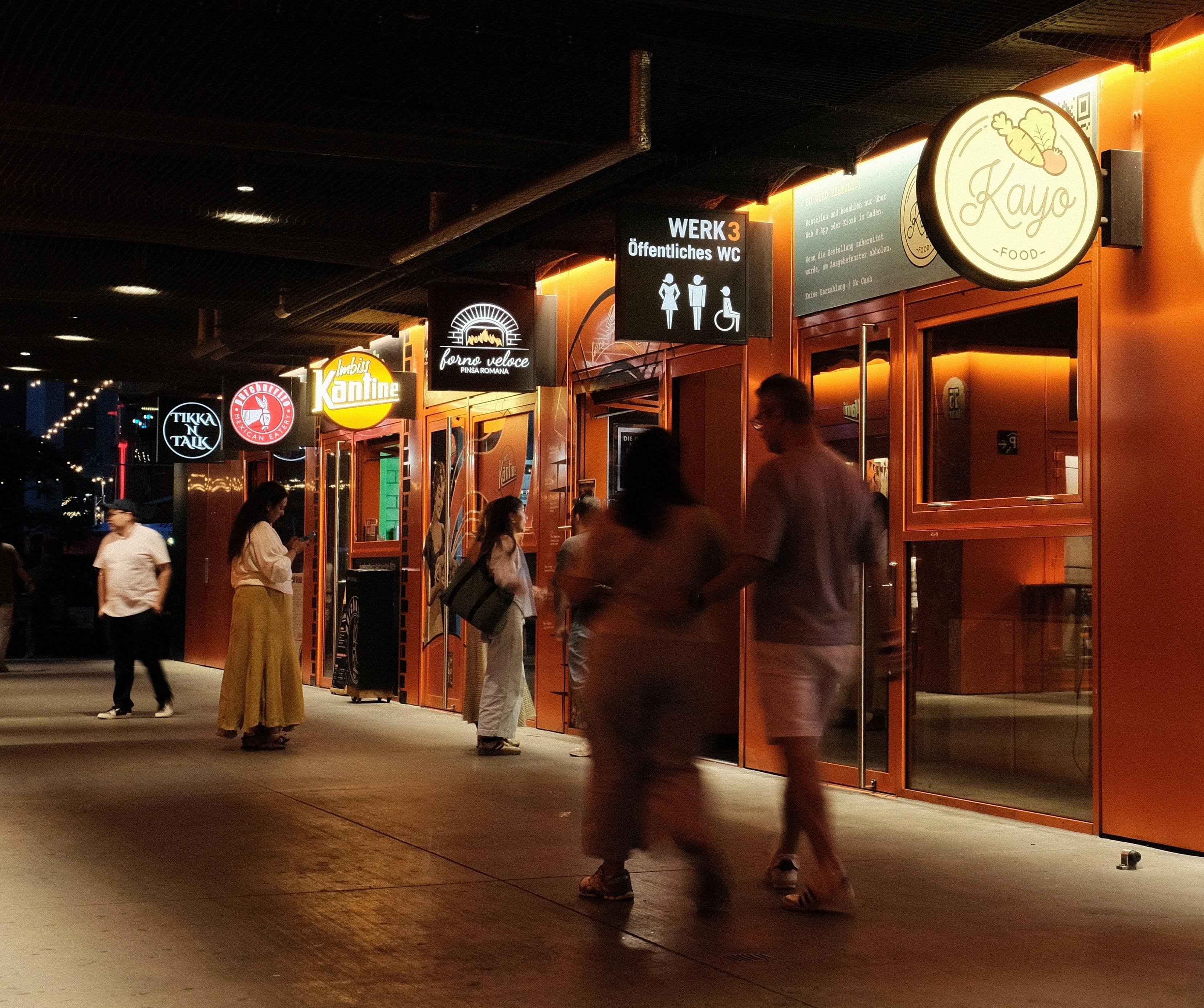 Nighttime scene in a pedestrian passage through a building; a row of orange storefronts with illuminated restaurant signs and a restroom sign reading “WERK 3 Öffentliches WC”; several pedestrians walk past, some blurred from motion.