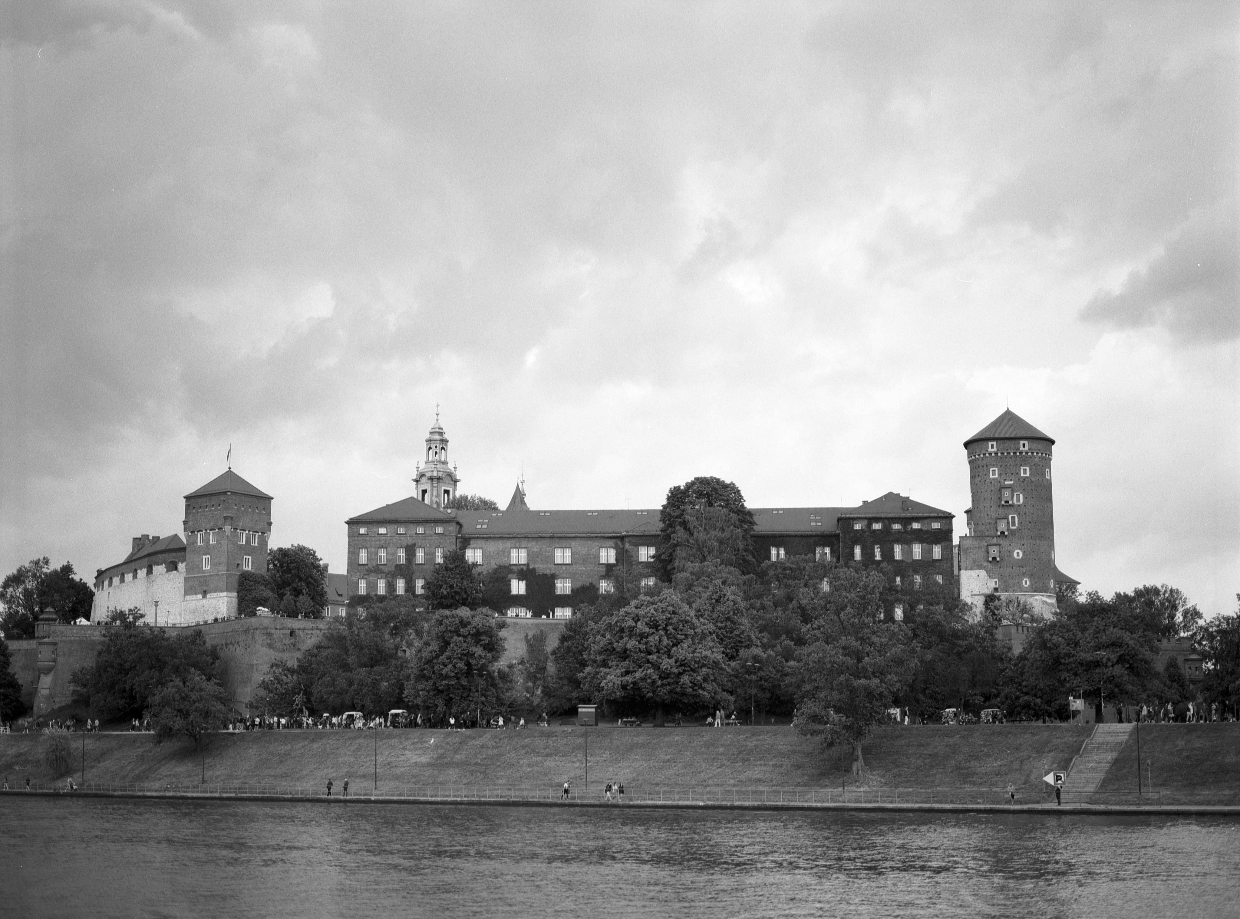 Historic castle (Wawel) with multiple towers and spires, situated on a hill with dense trees. A river flows in the foreground, and several people are visible on a path along the water. The sky is overcast with thick clouds.