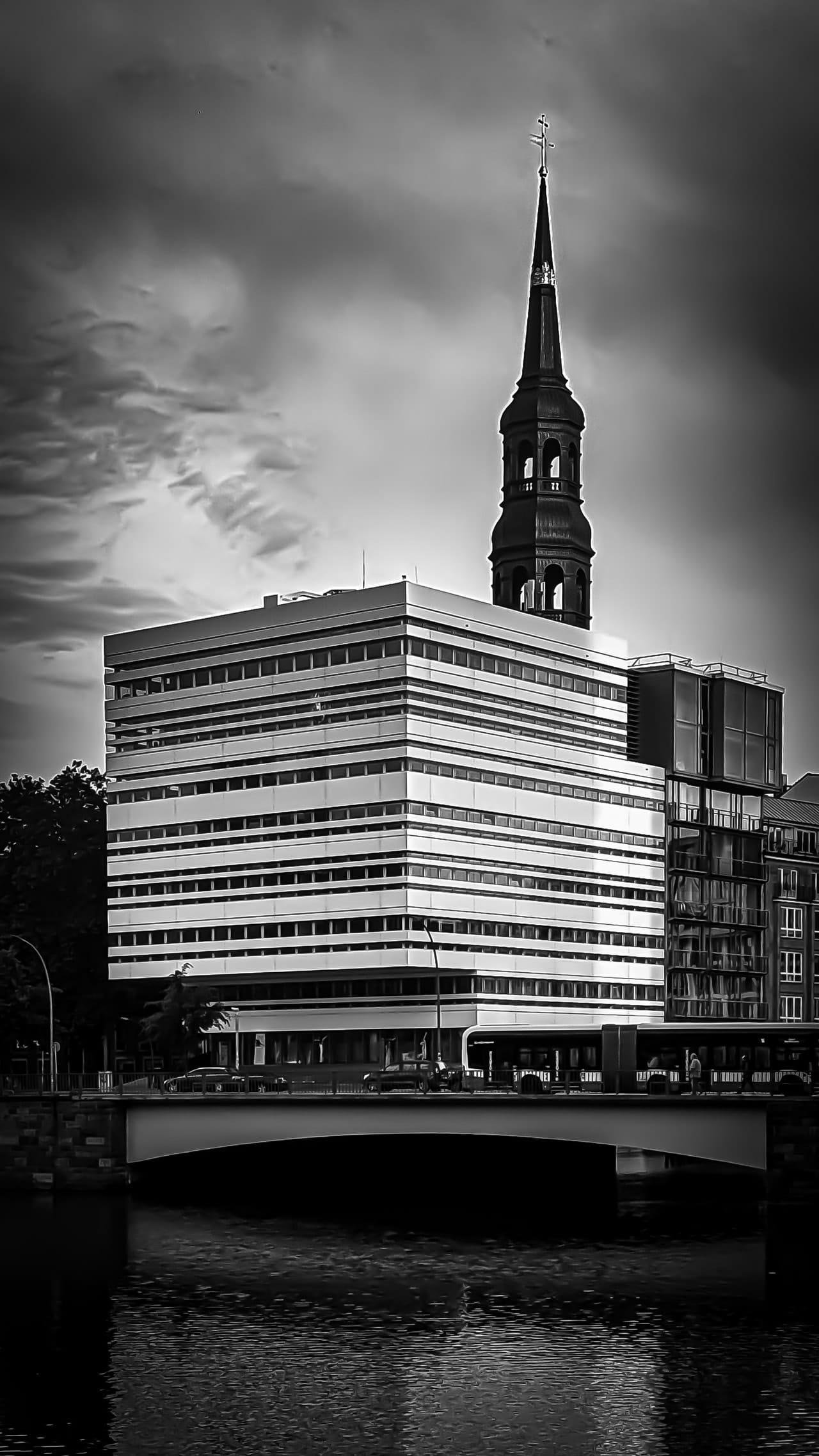 Black-and-white cityscape of a modern, striped office building beside a river, with a bridge and a bus in the foreground and a tall church spire topped with a cross rising behind the building under a cloudy sky.