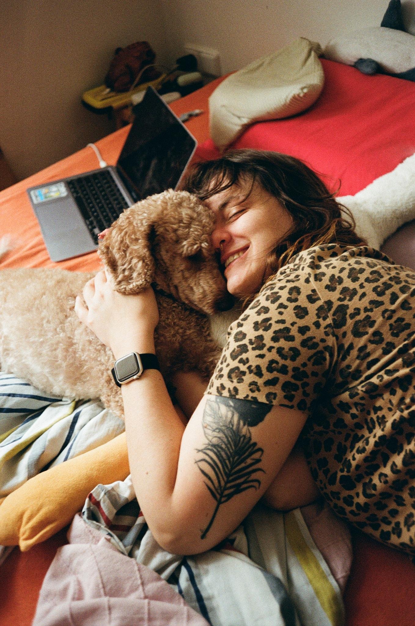Person lying on a bed hugs a small curly-haired dog; a laptop sits open behind them on the red bedsheet, with pillows and a patterned blanket in the foreground.