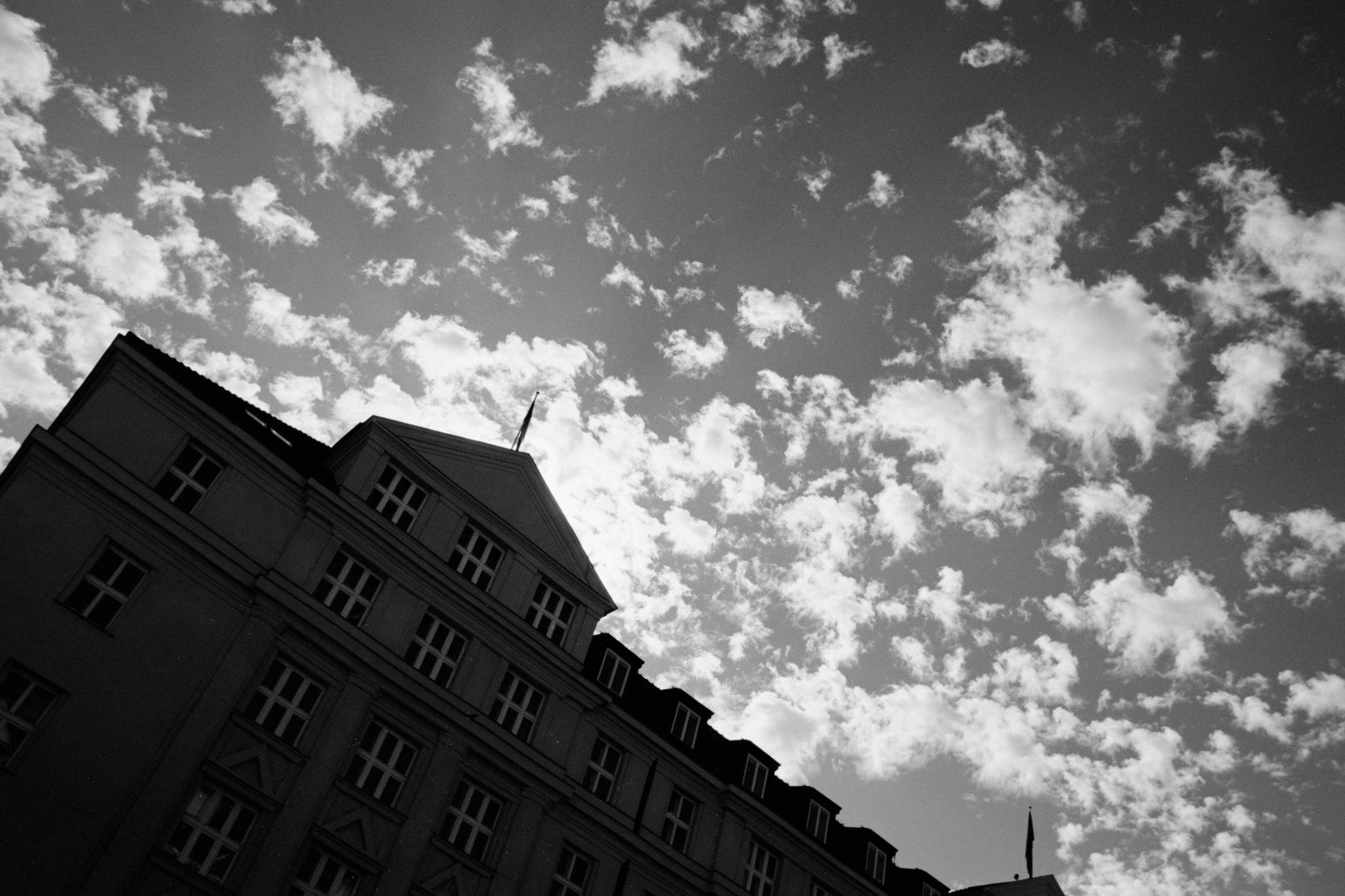 Low-angle black-and-white view of a multi-story building corner with rows of windows and dormers, set against a sky filled with scattered clouds.