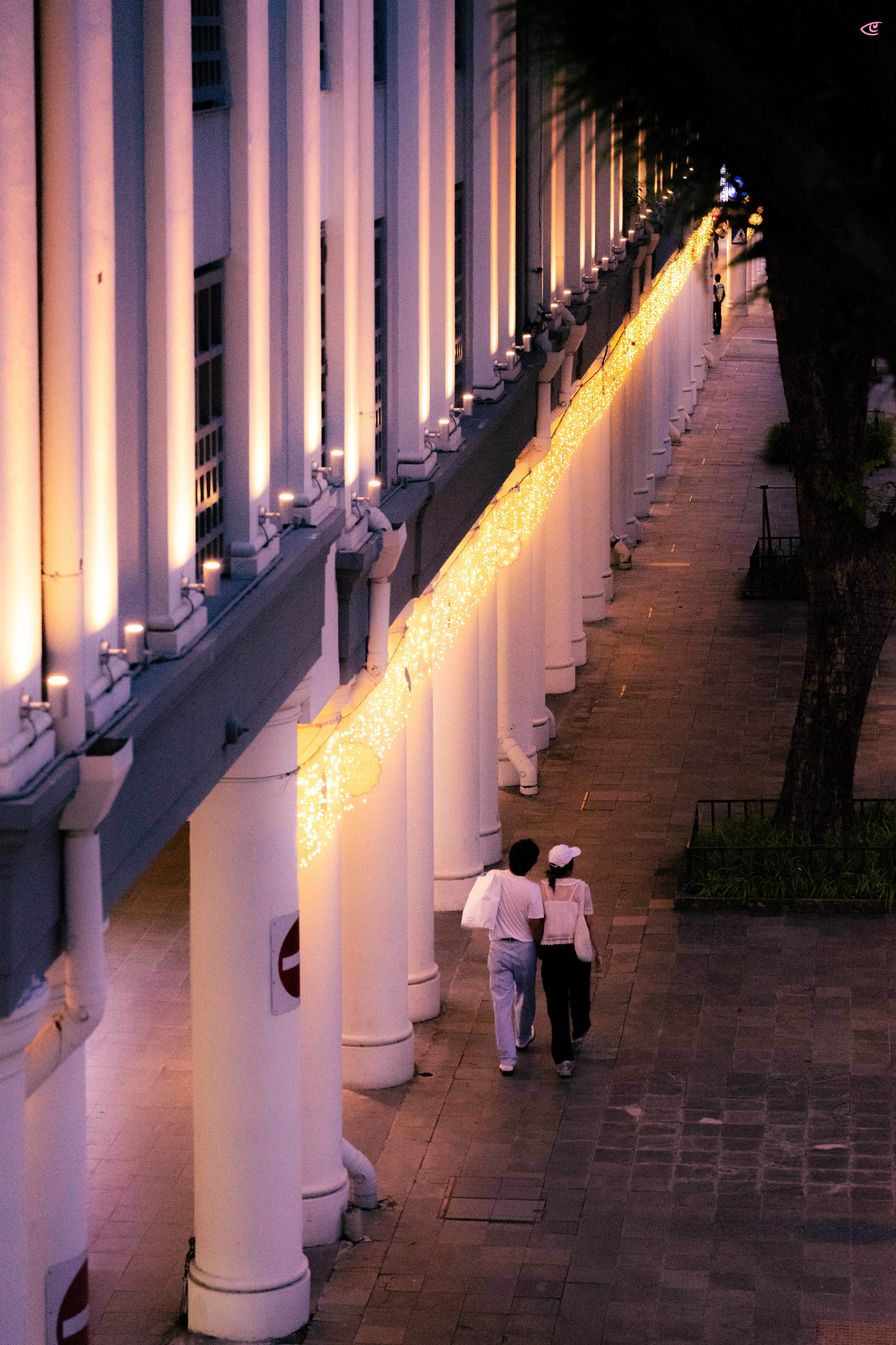 Long colonnade of white pillars along a sidewalk at dusk, lit by a continuous string of warm fairy lights; two people walk away in the foreground while trees line the right side.