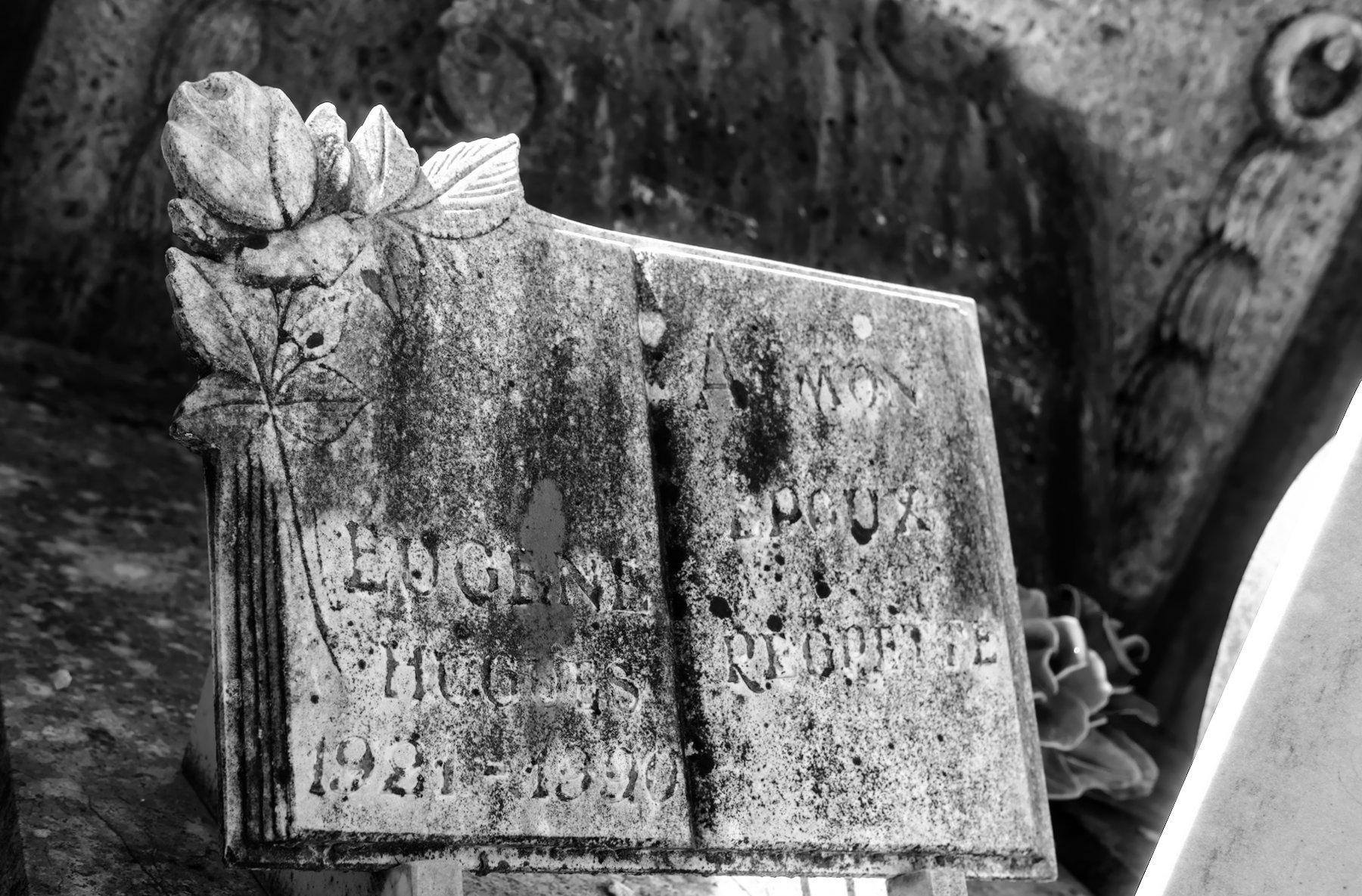 Photo horizontale en noir et blanc. Gros plan sur une plaque commémorative sur une tombe, en forme de livre avec le coin supérieur gauche décoré d’une fleur et dont les écritures ont été fortement abimées par le temps