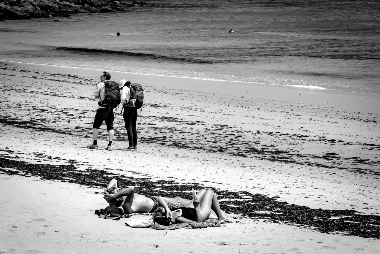 Two people sunbathing on towels in the foreground on a sandy beach. In the middle ground, two hikers with backpacks stand facing the ocean. A swimmer is visible far in the water. The beach has patches of seaweed, and rocky terrain borders the shoreline. The image is in black and white.
