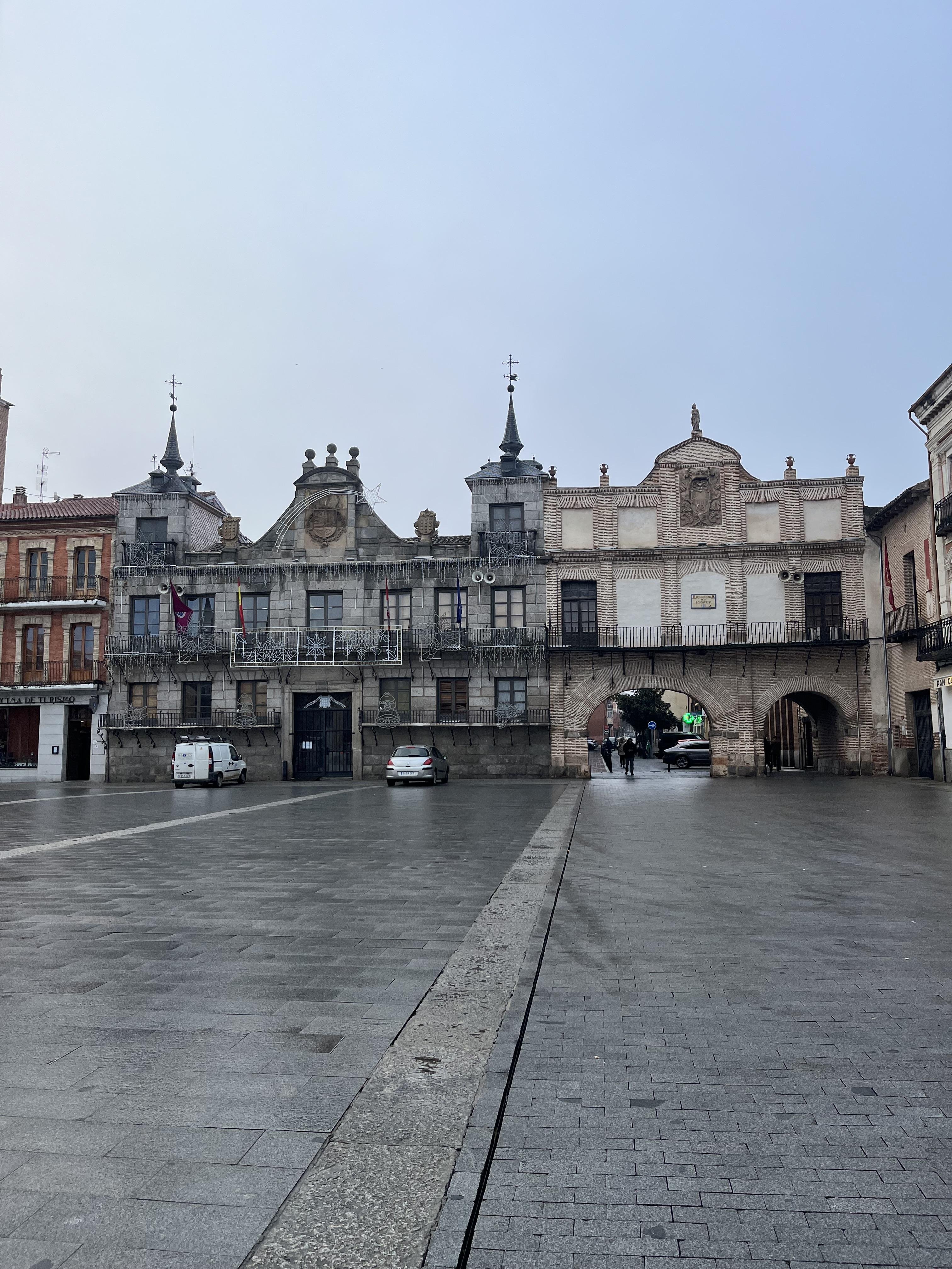 The image shows a historic building with an ornate facade, featuring a series of arches and decorative elements. It seems to be located in a plaza or open square, with cobblestone or paved ground. There are a few vehicles parked nearby, and some people are walking under the arches. The architecture is likely European, possibly Spanish, given the style and design features.