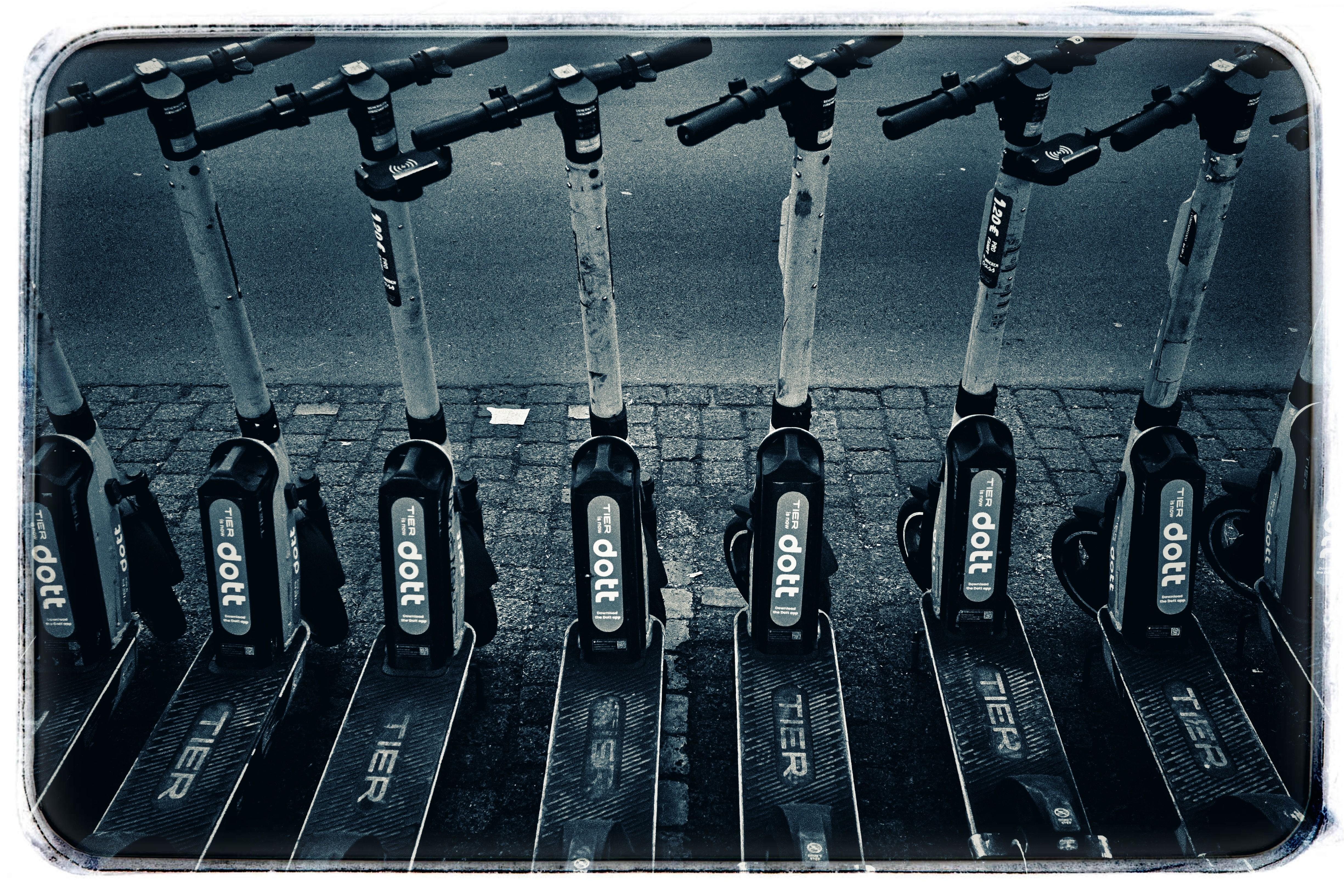 Row of parked rental e-scooters lined up side by side on a cobblestone sidewalk, with handlebars and stems visible above black decks near the curb. ### 
Eine Reihe geparkter Miet-E-Scooter steht nebeneinander auf einem Kopfsteinpflaster-Gehweg, Lenker und Lenkstangen sind über den schwarzen Trittbrettern nahe dem Bordstein sichtbar.