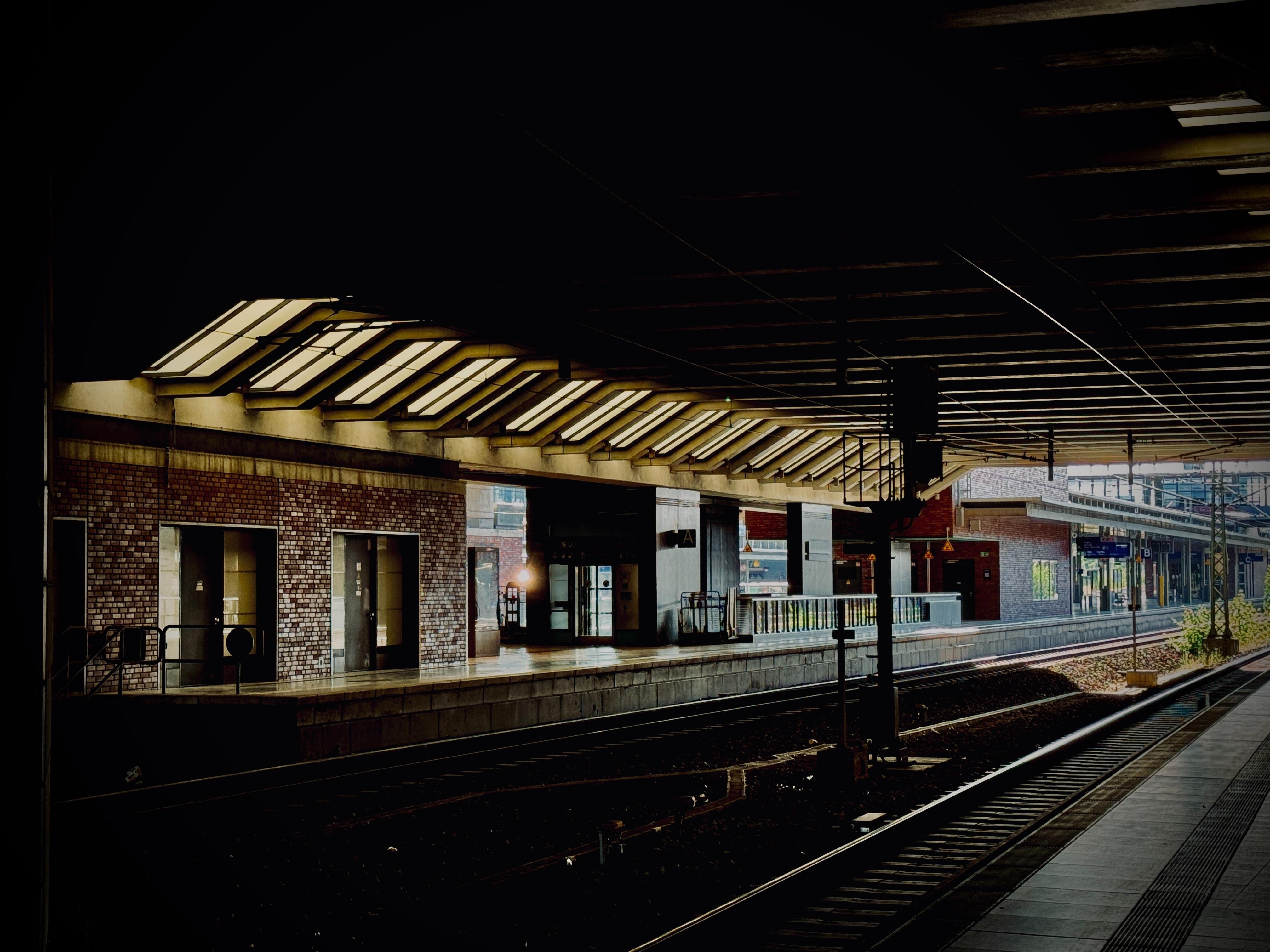 A train station platform with two train tracks. The platform has a brick facade with large windows and a canopy with translucent panels.