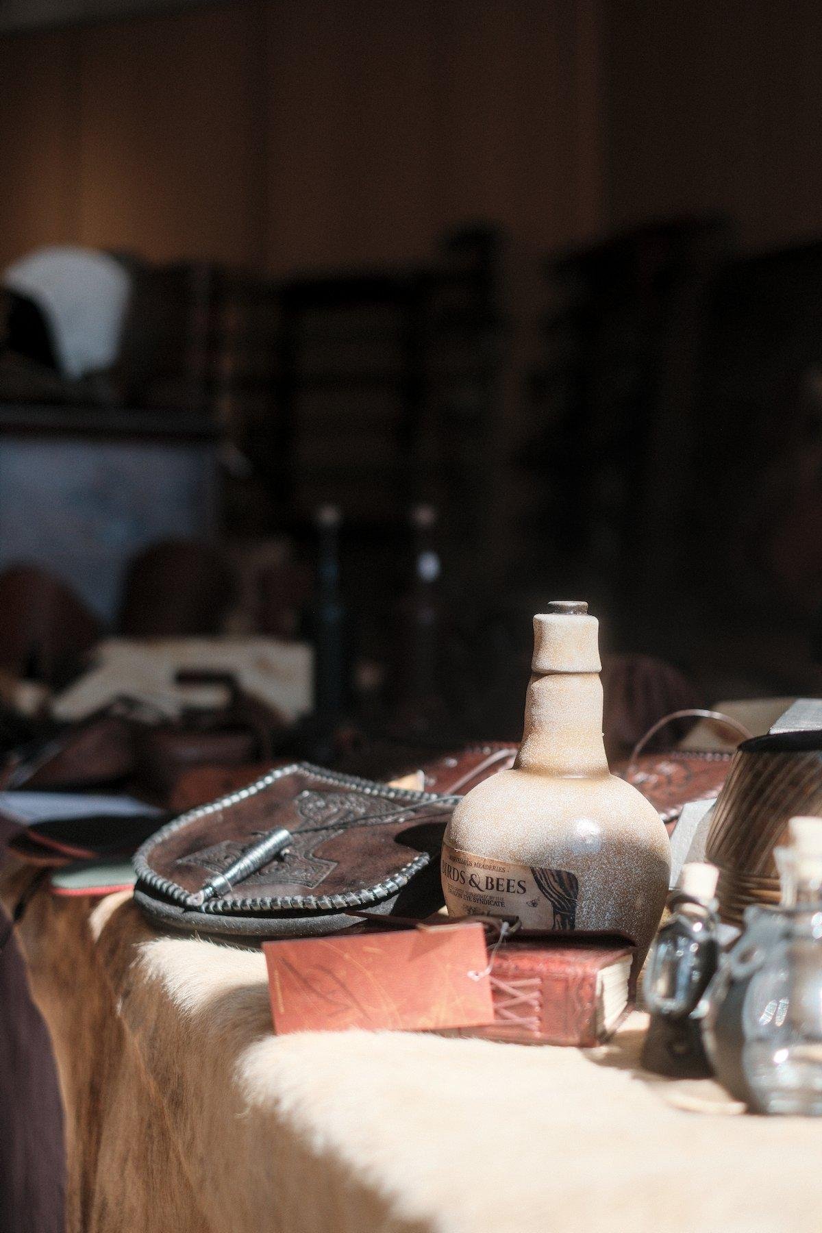 Photo verticale. Sur une table couverte d’une épaisse nappe beige, des marchandises de type heroic fantasy sont disposées à la vente. Une petite bouteille en terre au corps sphérique, une sacoche en cuir, des fioles en verre… le tout éclairé par le soleil entrant par une fenêtre derrière la caméra