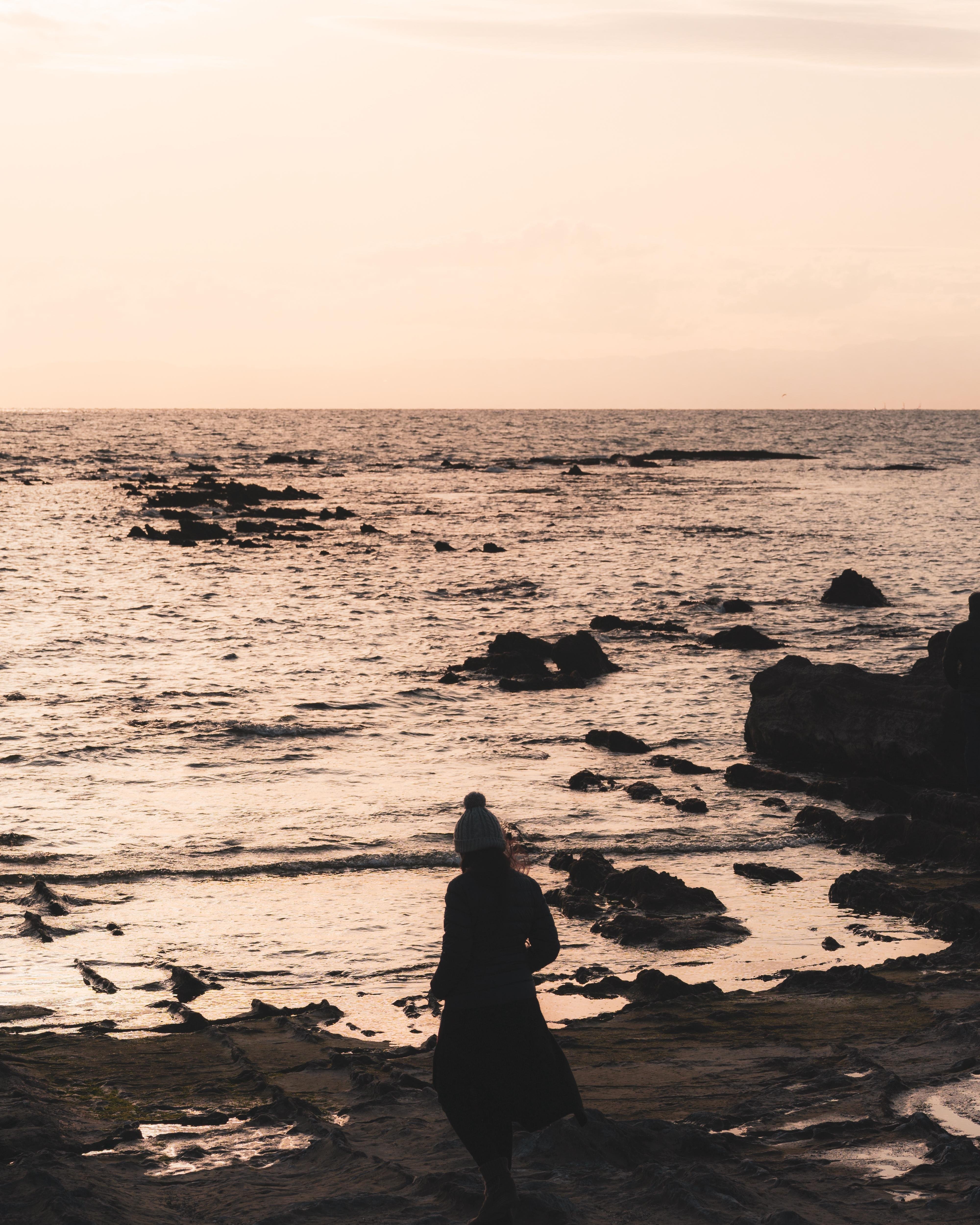 The image shows a person standing by the seashore at sunset. The ocean is visible, with waves gently hitting the rocky shoreline. The sky has a warm, golden hue typical of a sunset. The person appears to be wearing a coat and a hat, possibly due to cool weather. The overall atmosphere is calm and serene.