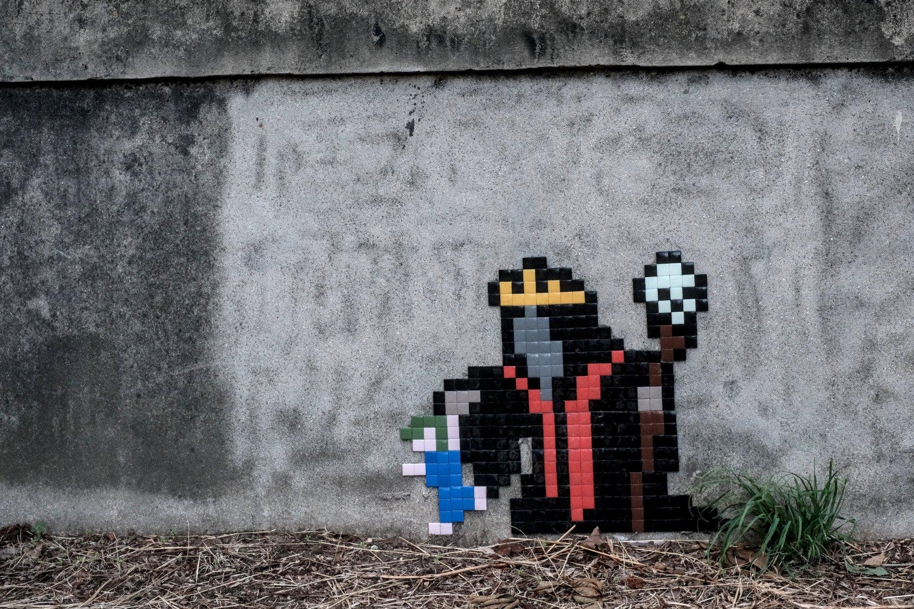 Photo horizontale. Sur un muret, au ras du sol, un pixelart représentant un personnage avec une couronne dorée, un robe noir et rouge, tenant un bâton surmonté d’un crâne de la main gauche, et un lemming de la main droite