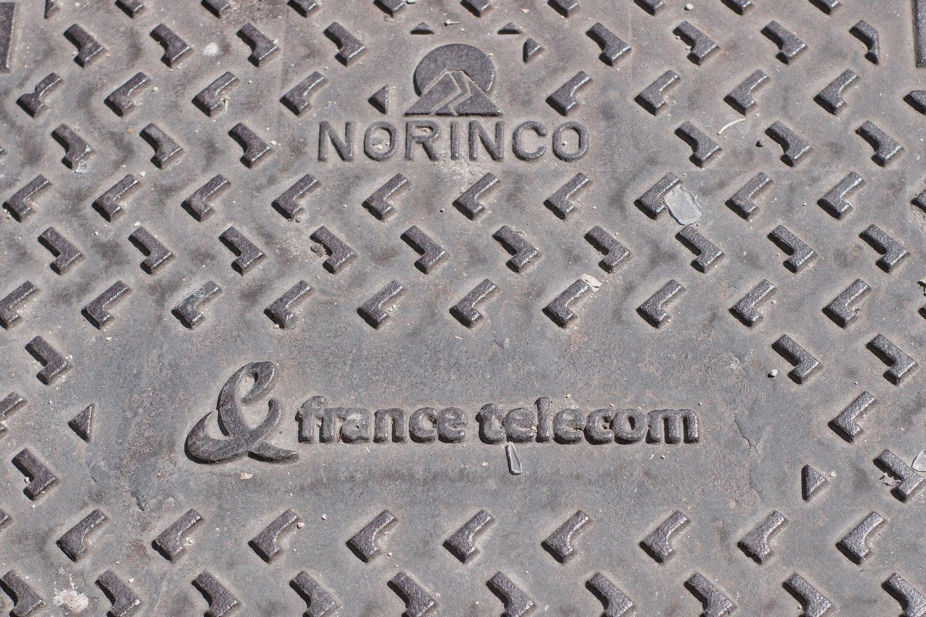 Photo horizontale. Gros plan sur une plaque en acier, une trappe de service marquée NORINCO et France Telecom