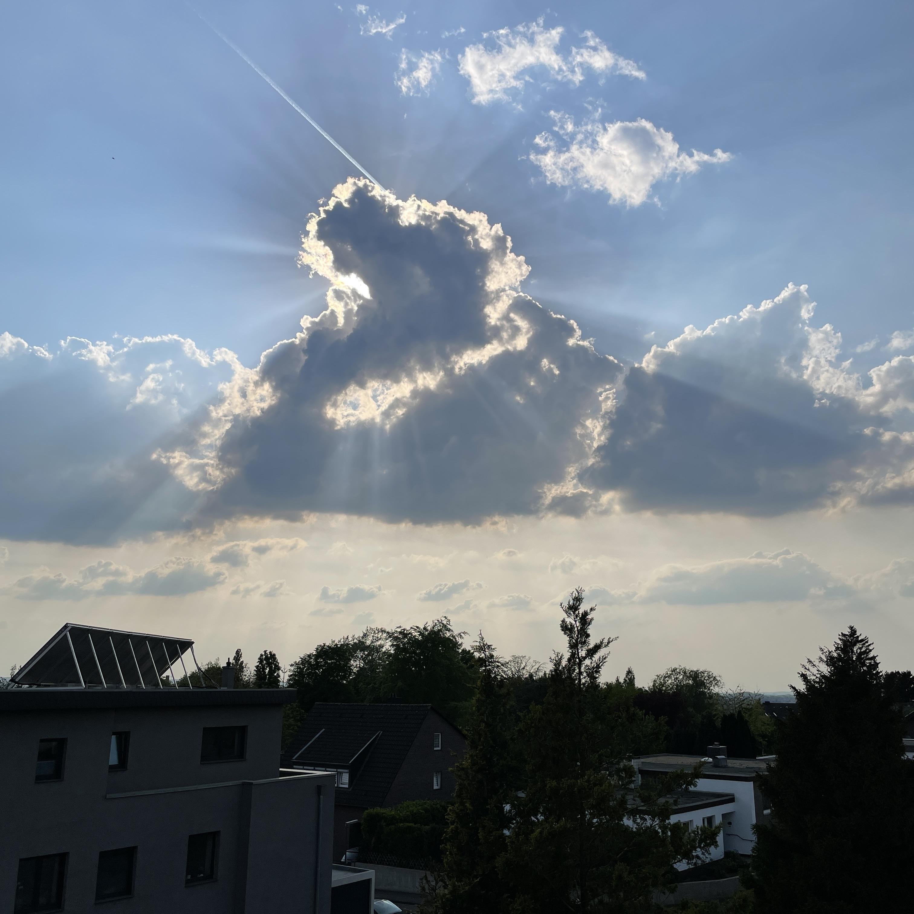 Das Bild zeigt einen Himmel mit auffälligen Wolkenformationen, durch die die Sonne scheint und strahlenförmige Lichtkegel erzeugt.  Im Vordergrund sind Dächer von Häusern und Bäume zu sehen. Der Kontrast zwischen dem hellen, von der Sonne durchbrochenen Himmel und den dunklen Gebäuden und Bäumen ist markant. Ein Flugzeugkondensstreifen ist ebenfalls sichtbar.

