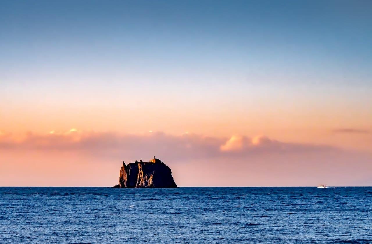 Small rocky island on the horizon in a calm ocean at sunset, with pink clouds in the sky and a tiny boat at far right.