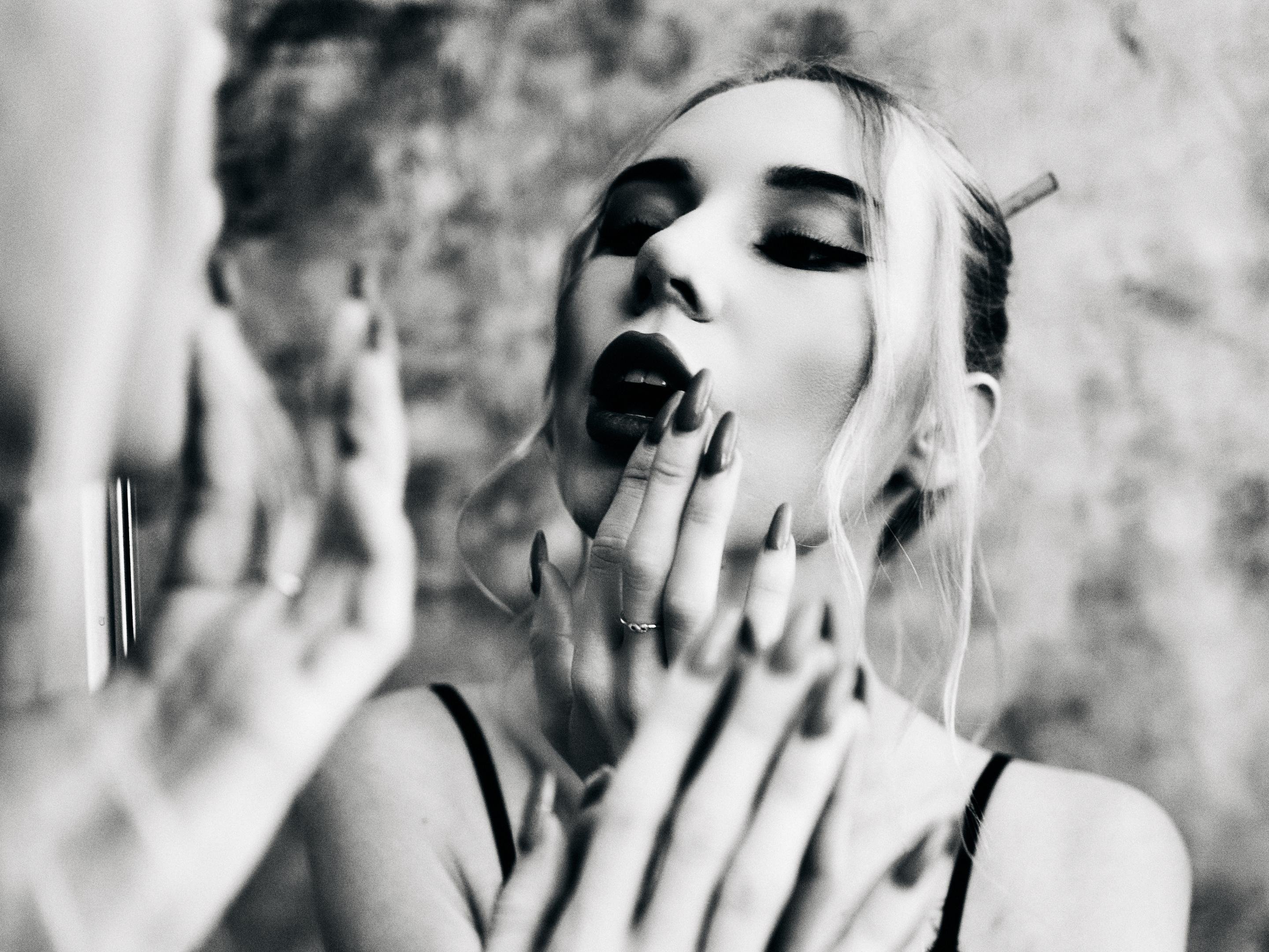 Black-and-white close-up of a woman facing a mirror, with her reflection blurred in the foreground; she wears dark eye makeup and lipstick, has light hair pulled back with loose strands, holds her hand with long pointed nails near her mouth, and a cigarette rests behind her ear.