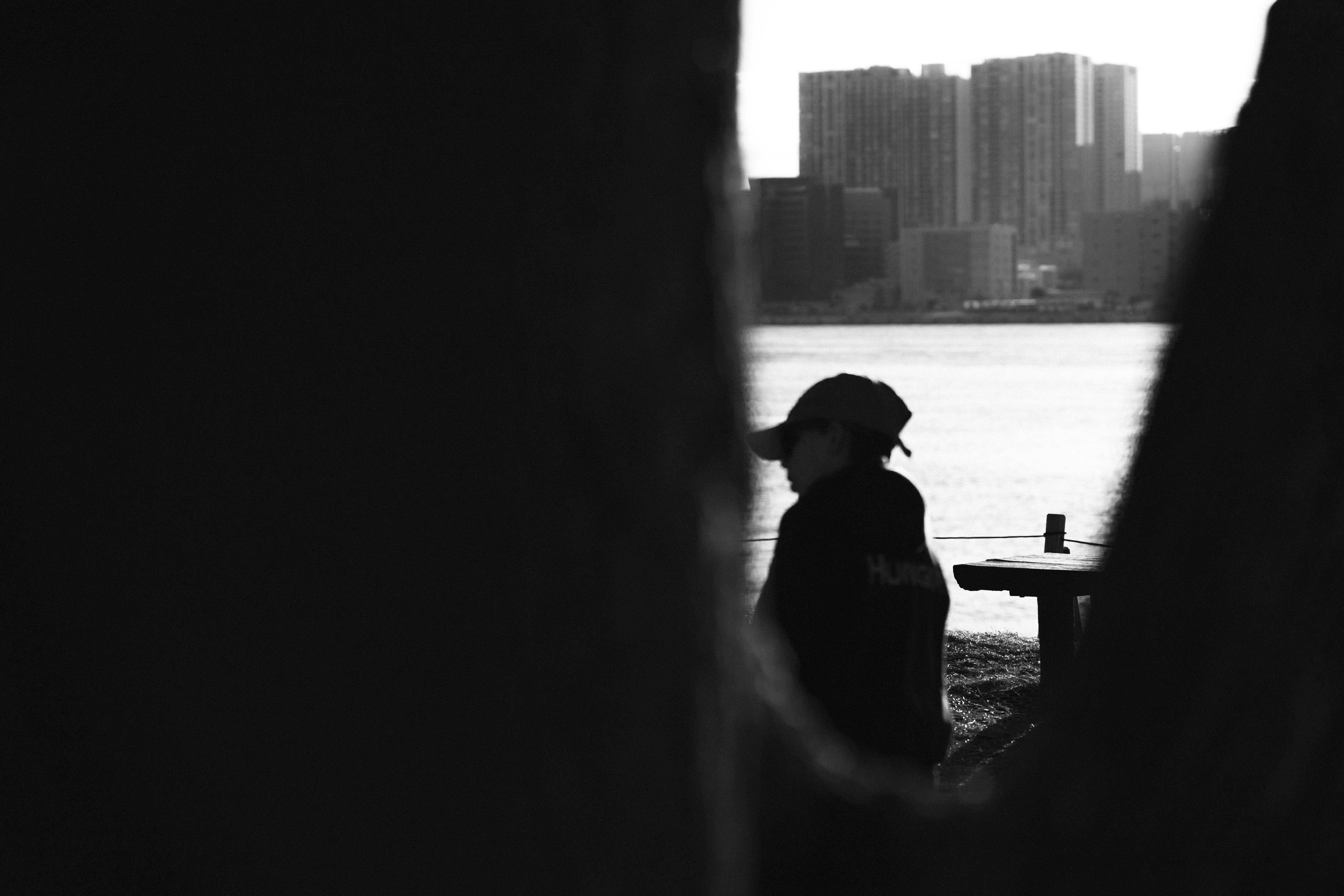 The image is in black and white and appears to show a person wearing a hat, possibly standing near a body of water. There are tall buildings in the background, suggesting an urban setting. The foreground is framed by tree trunks, creating a natural border. The image evokes a contemplative or serene mood.