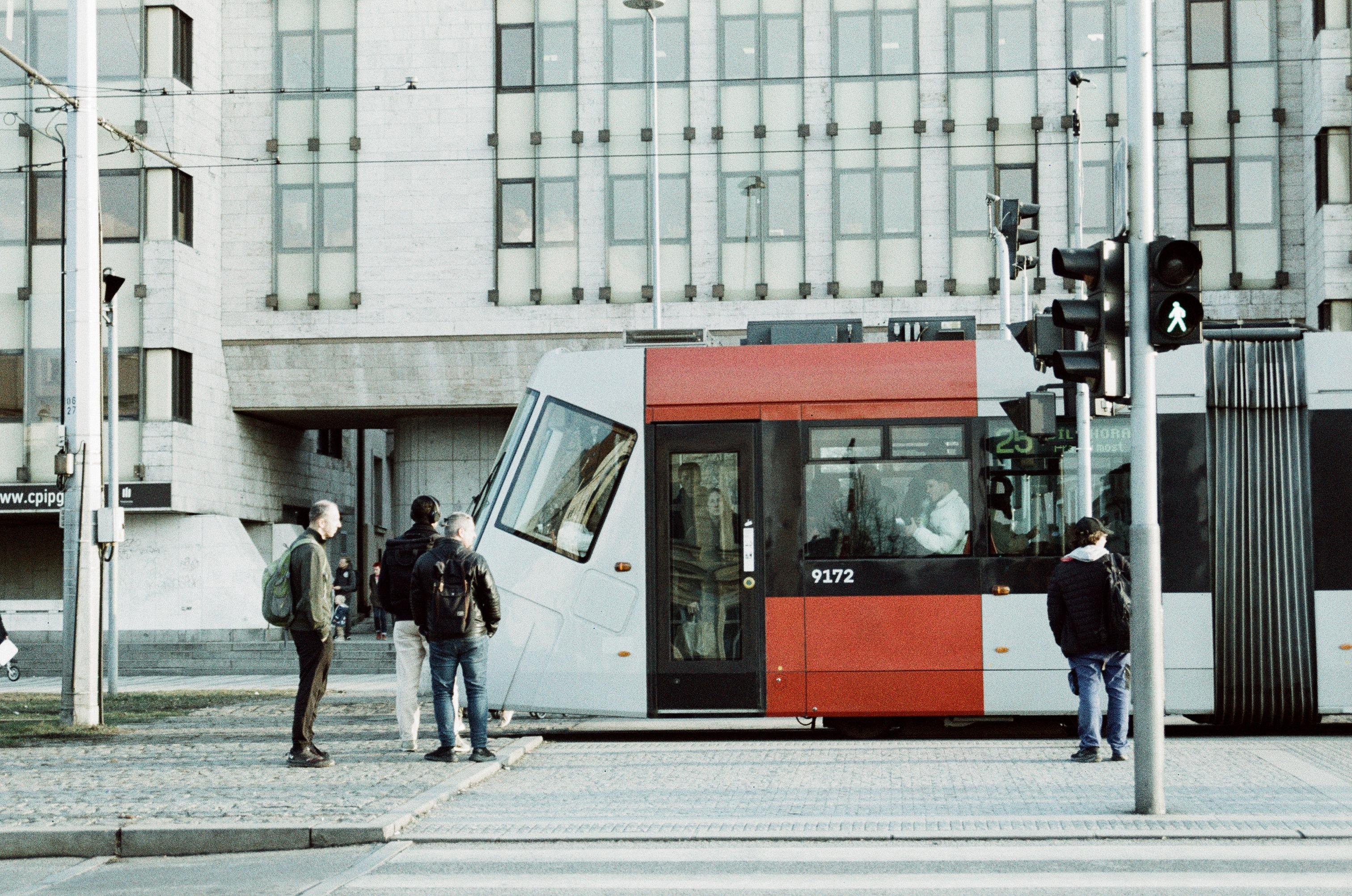 A muted-color photo of a rear end of a tram passing a pedestrian crossing with people bumbling about agaist a backdrop of a opressive modernist building